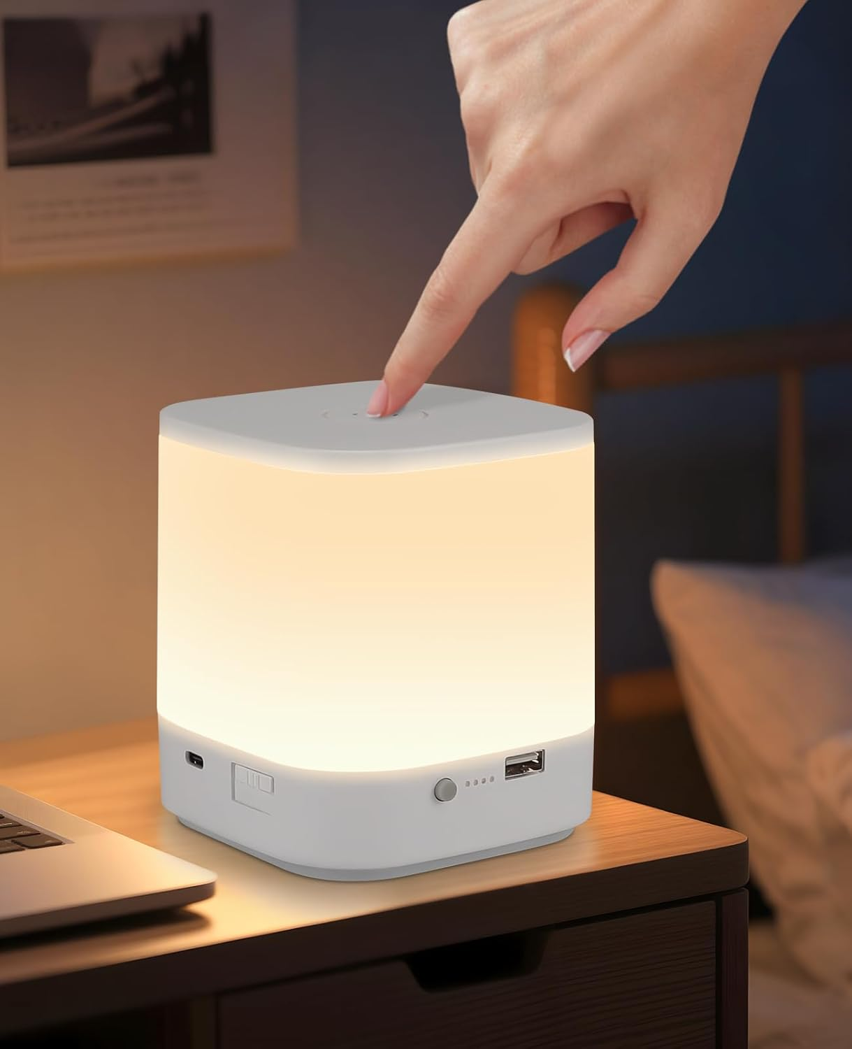 Biała lampa w kształcie kostki jest zapalona. Palec naciska przycisk u góry. Widoczny jest laptop.
