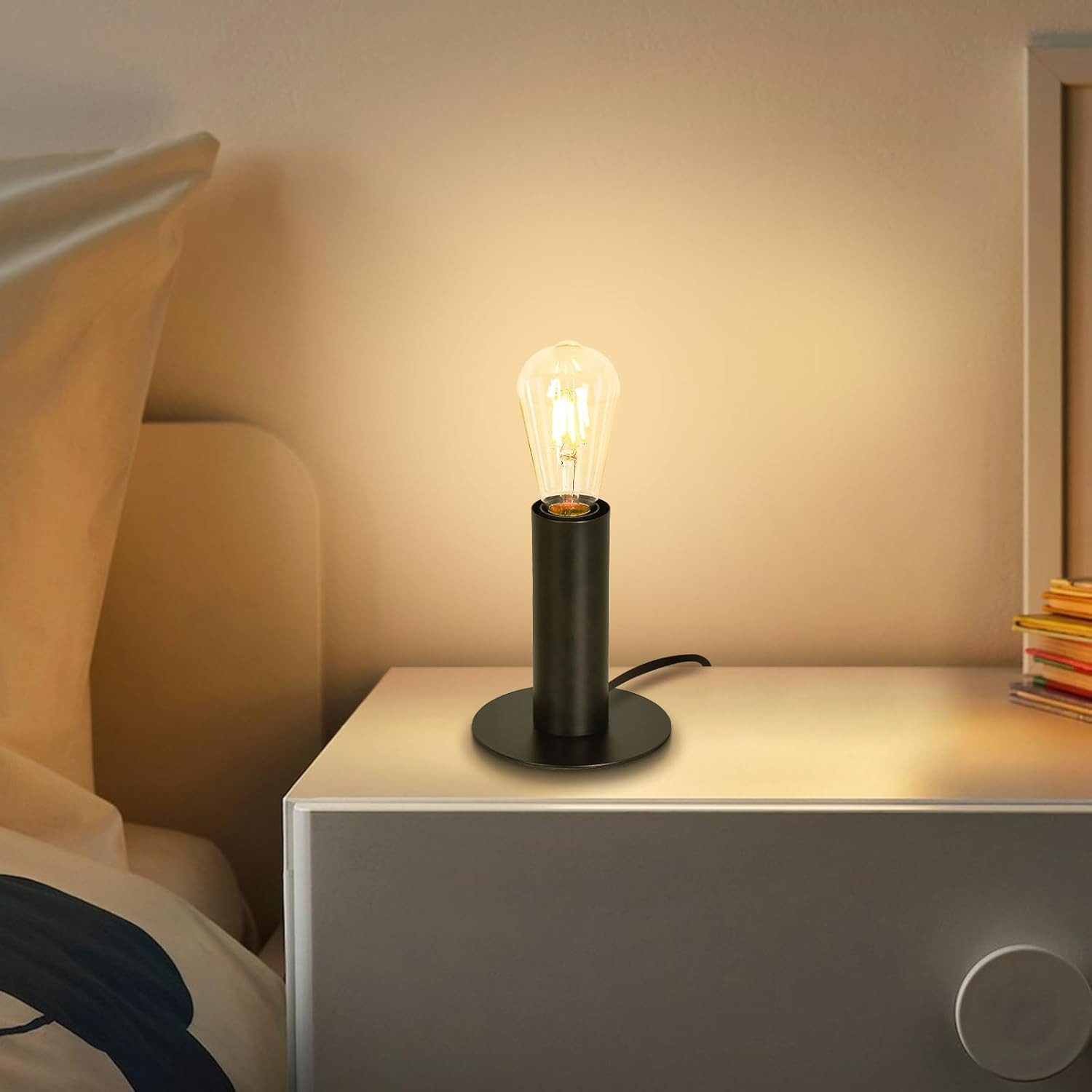 Czarna lampa z żarówką w stylu vintage na stoliku nocnym. Biała szuflada znajduje się pod lampą.