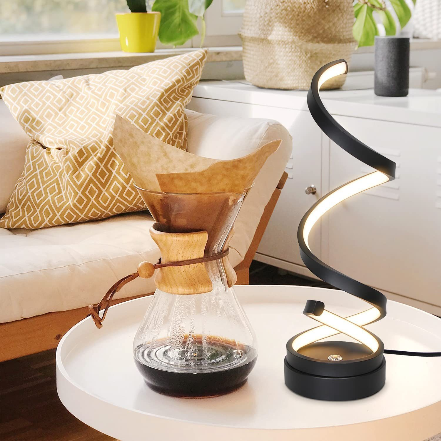 Kawiarka z kawą na białym stole, lampa spiralna i sofa z poduszkami w tle.
