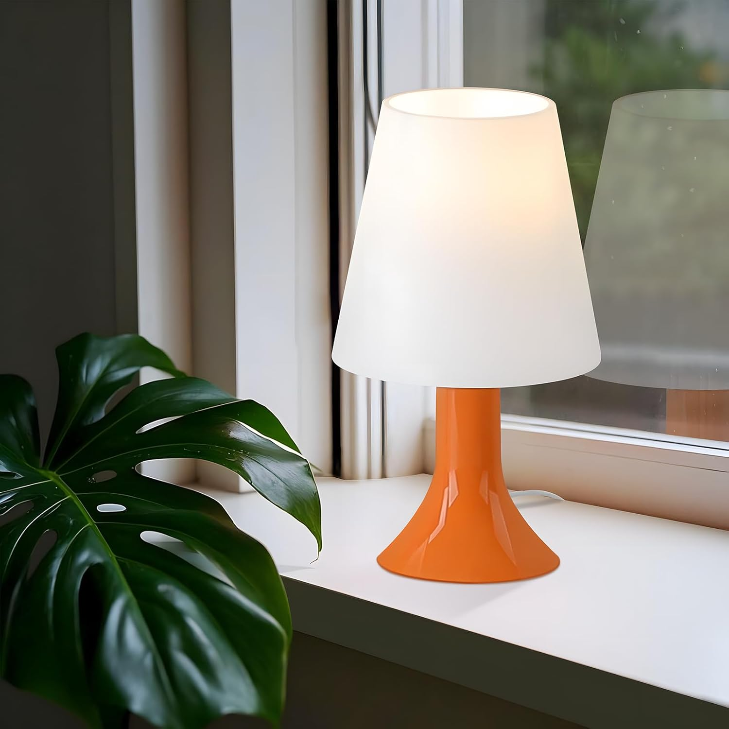 Pomarańczowa lampa na parapecie z dużym zielonym liściem na pierwszym planie.