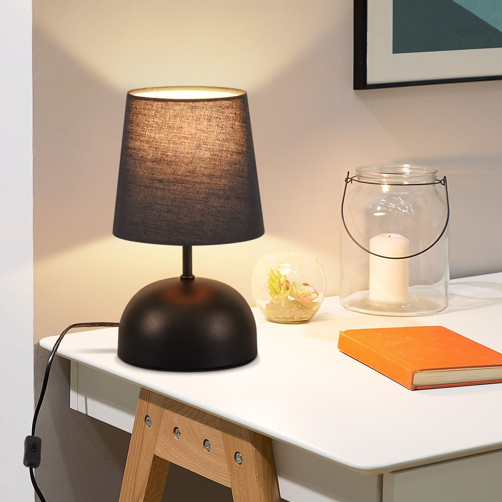 Czarna lampa stołowa z czarnym abażurem na białym stole. Włącznik po lewej.