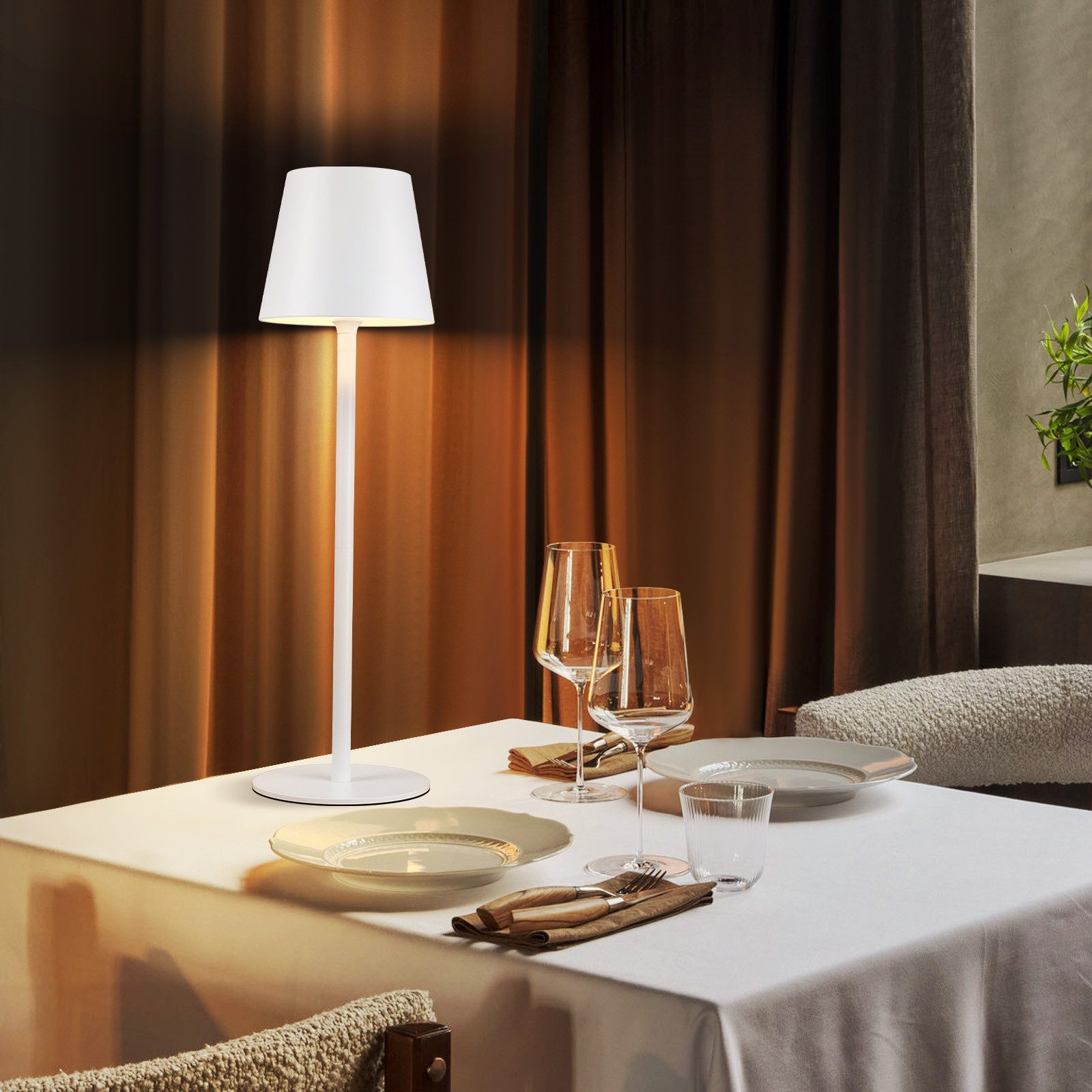 Biała lampa na stole. Na stole są szklanki i talerze. Tłem jest brązowa zasłona.
