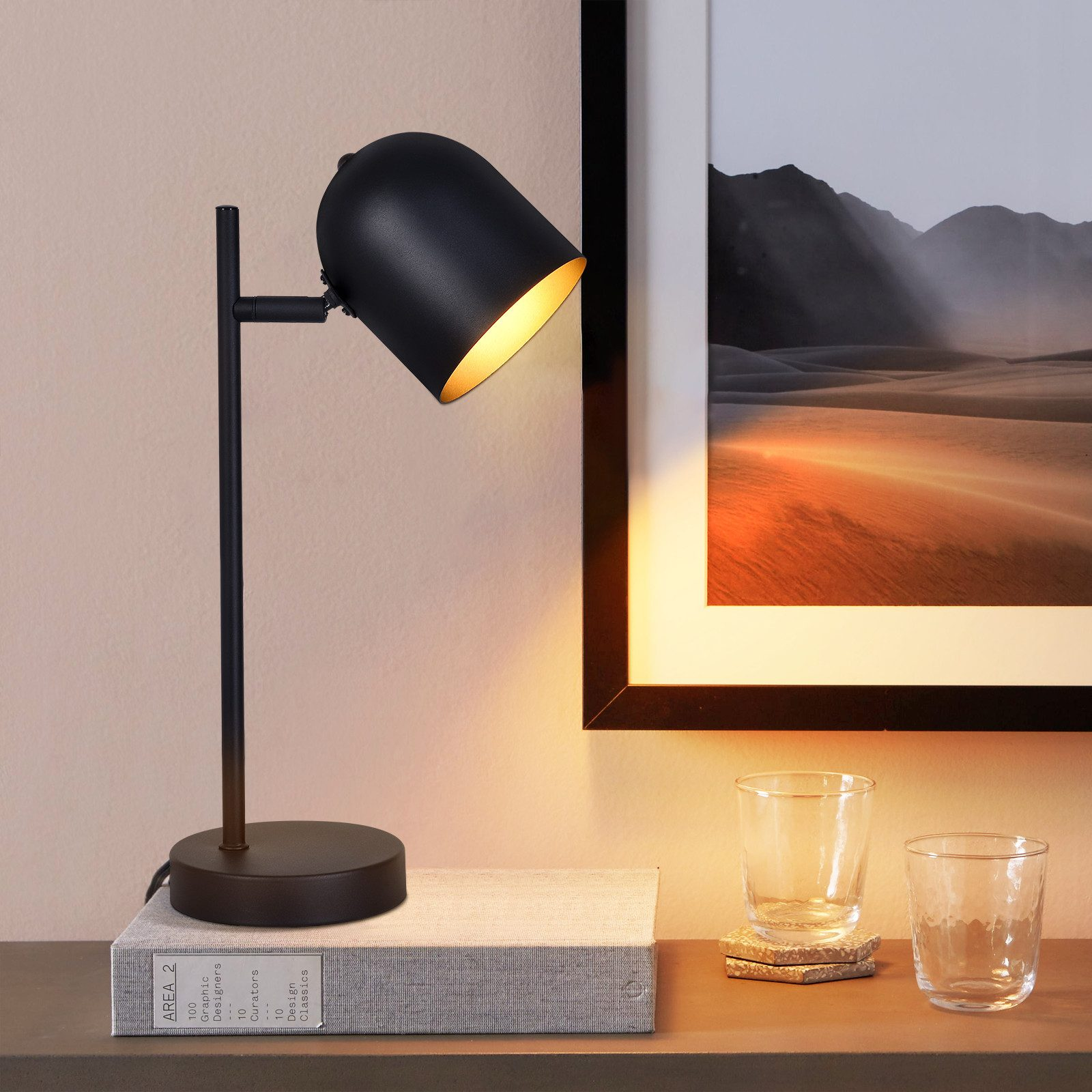 Czarna lampa biurkowa z zakrzywionym kloszem. Stoi na biurku z książkami i szklankami.