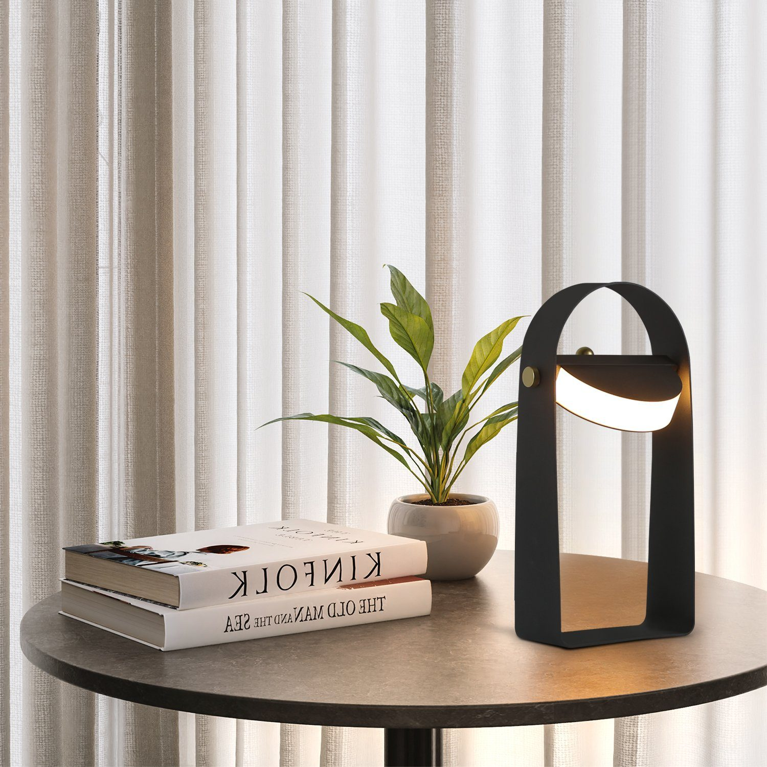 Czarna lampa z zakrzywioną ramą, rośliną i książkami na stole. Lampa ma światło w kształcie półksiężyca.