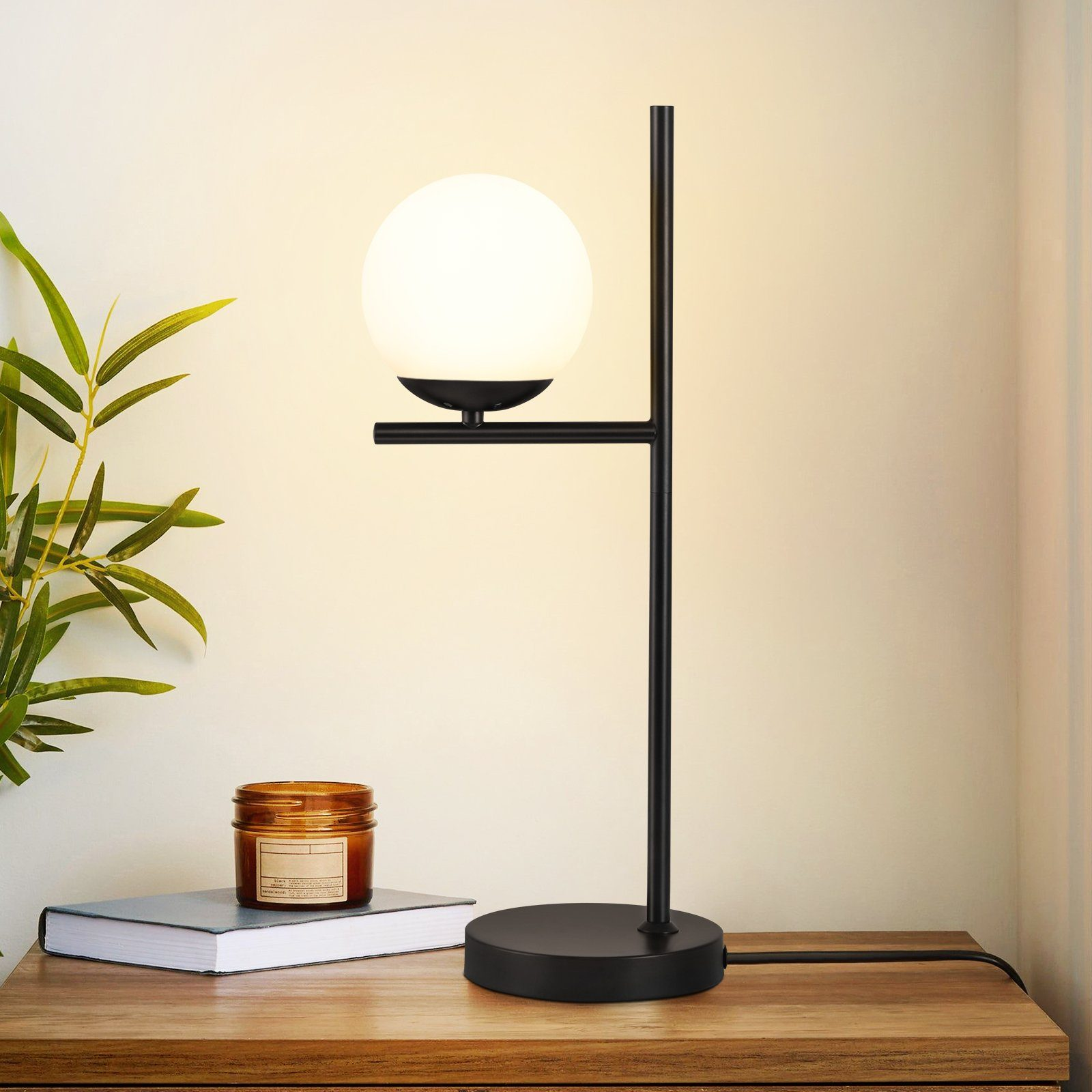 Czarna lampa z białym kloszem w kształcie kuli na czarnym stojaku, świeca i książka na drewnianej powierzchni.