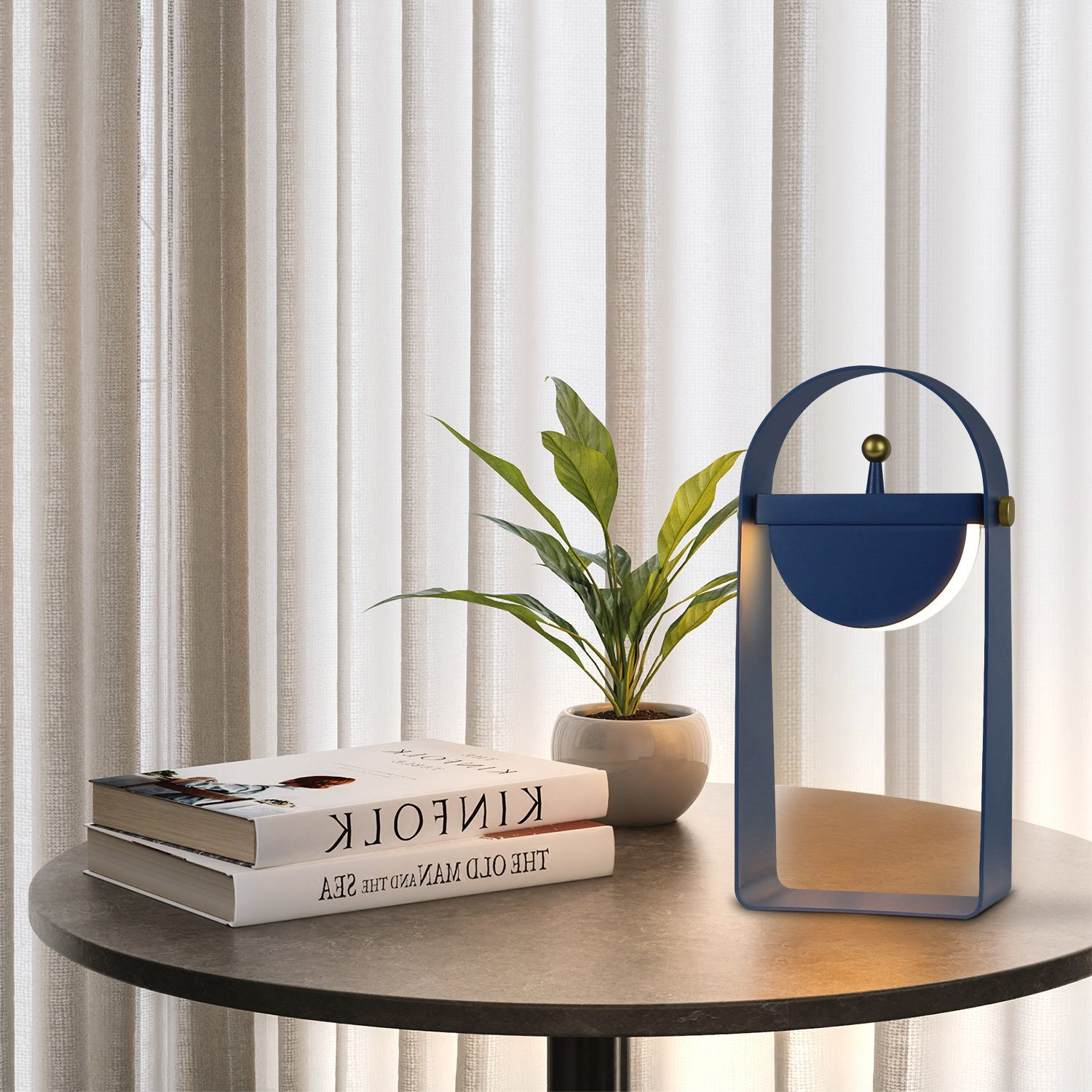 Lampa, książki i roślina na okrągłym stole, z zasłoną w tle.