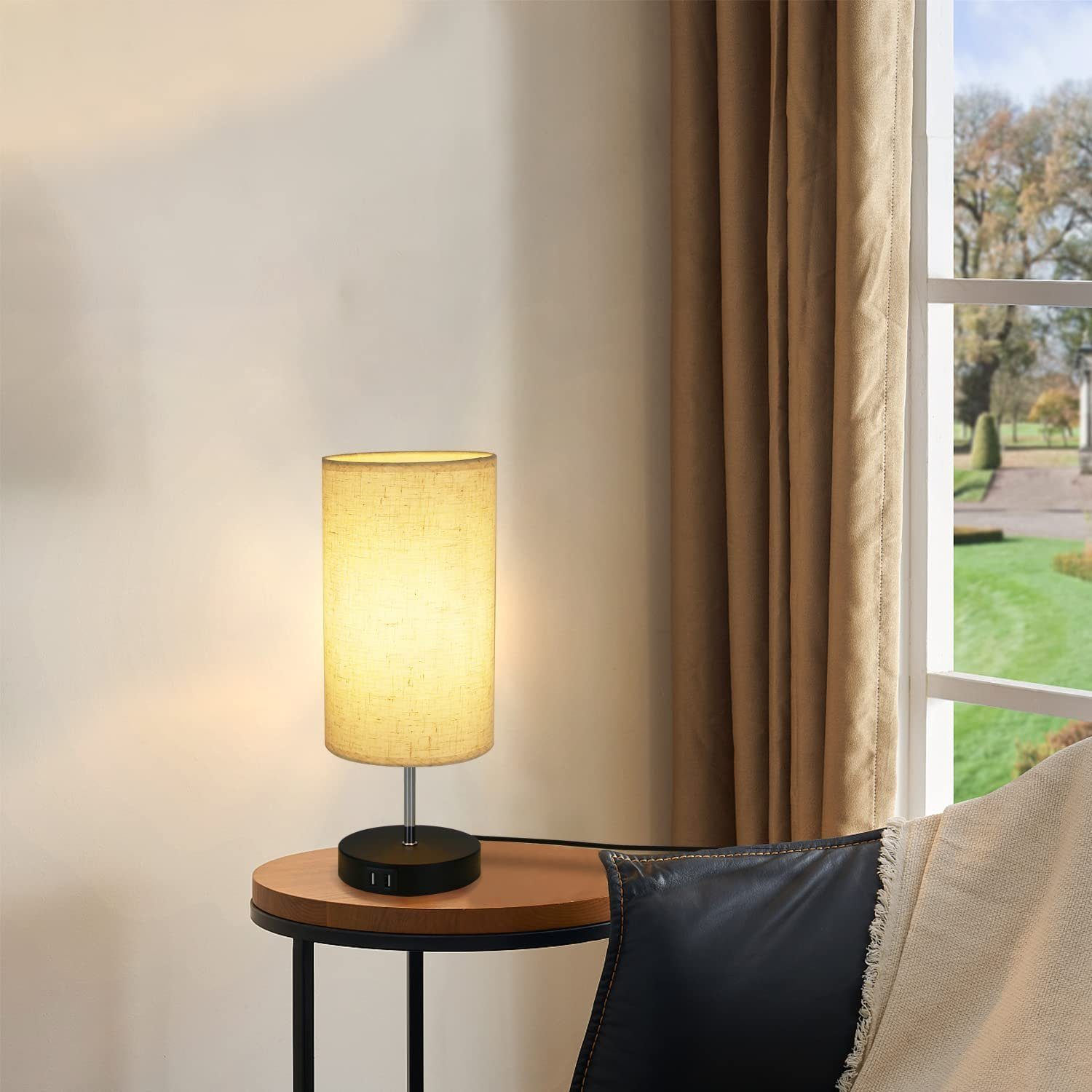 Lampa stołowa z beżowym kloszem i czarną podstawą stoi na drewnianym stole obok okna z widokiem.