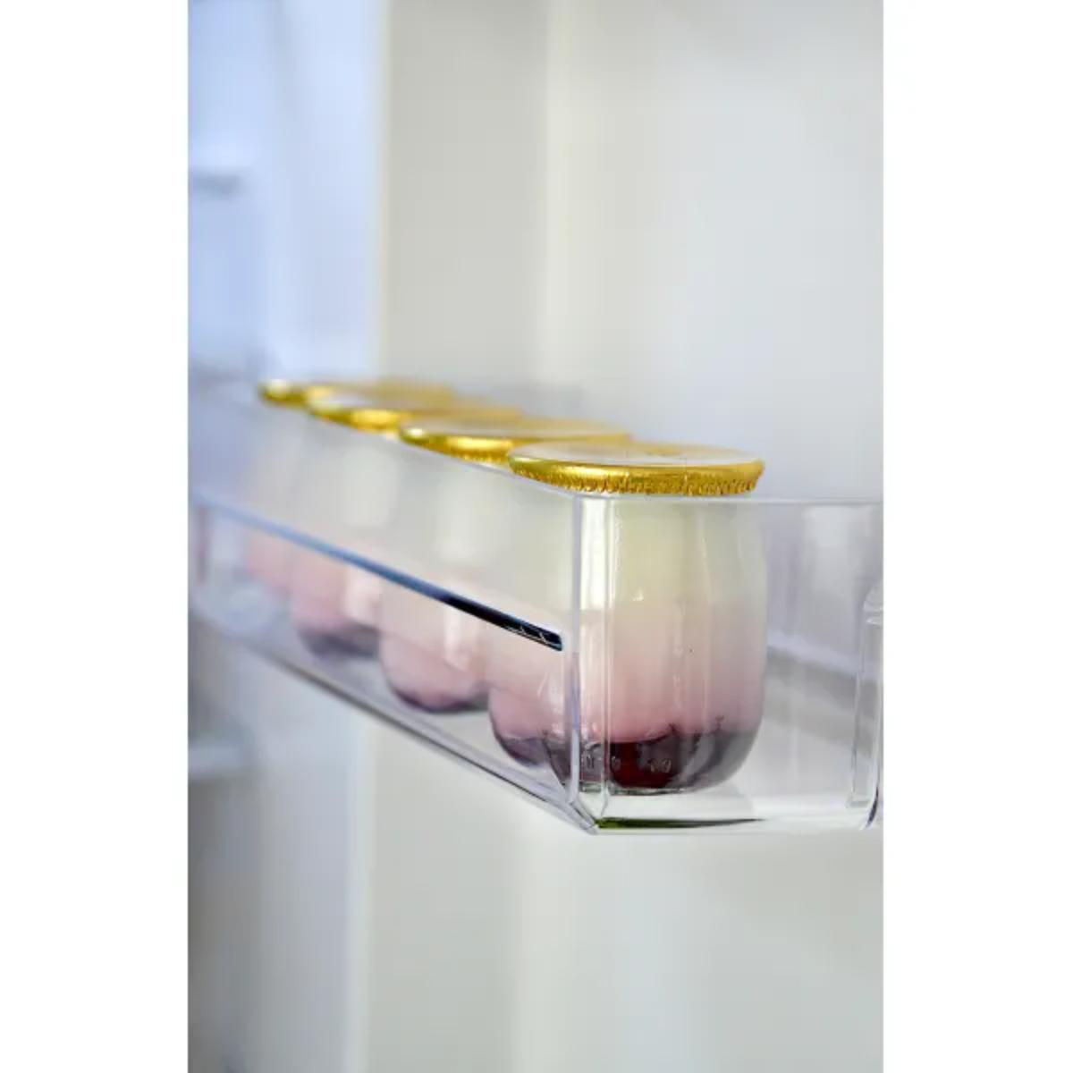 Półka w lodówce, na której znajdują się słoiczki różowego i białego jogurtu ze złotymi wieczkami. Tło jest białe.