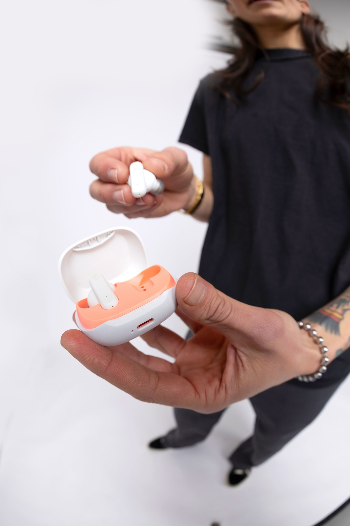 Osoba trzyma białe i koralowe słuchawki w białym etui. Tło jest białe. Osoba ma na sobie czerń.