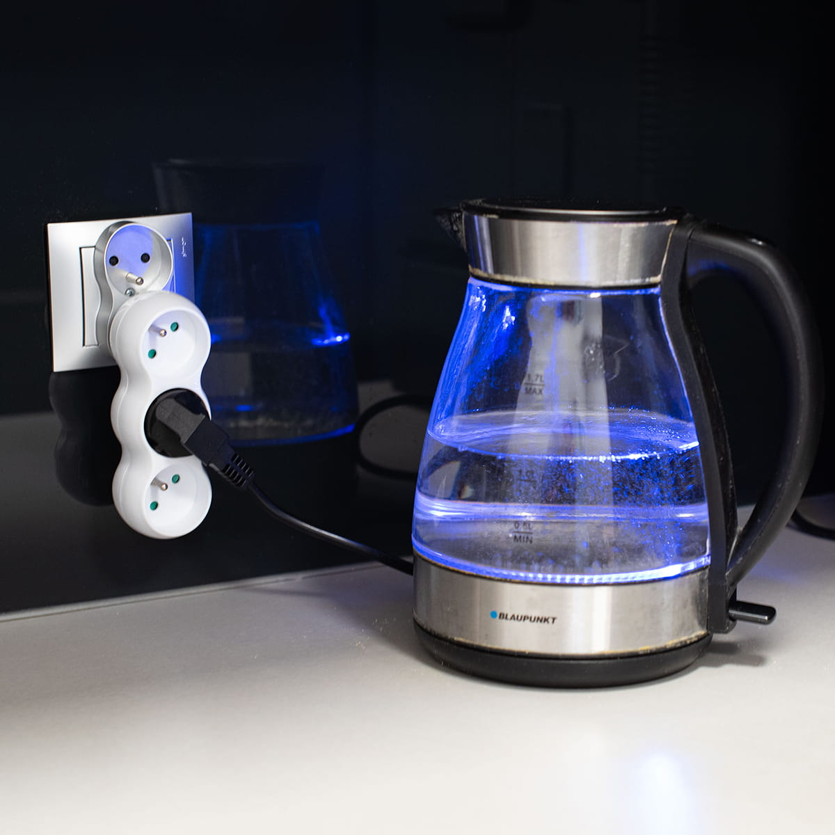 Biały adapter z wtyczką i czajnik z niebieskim światłem. Czarne tło.