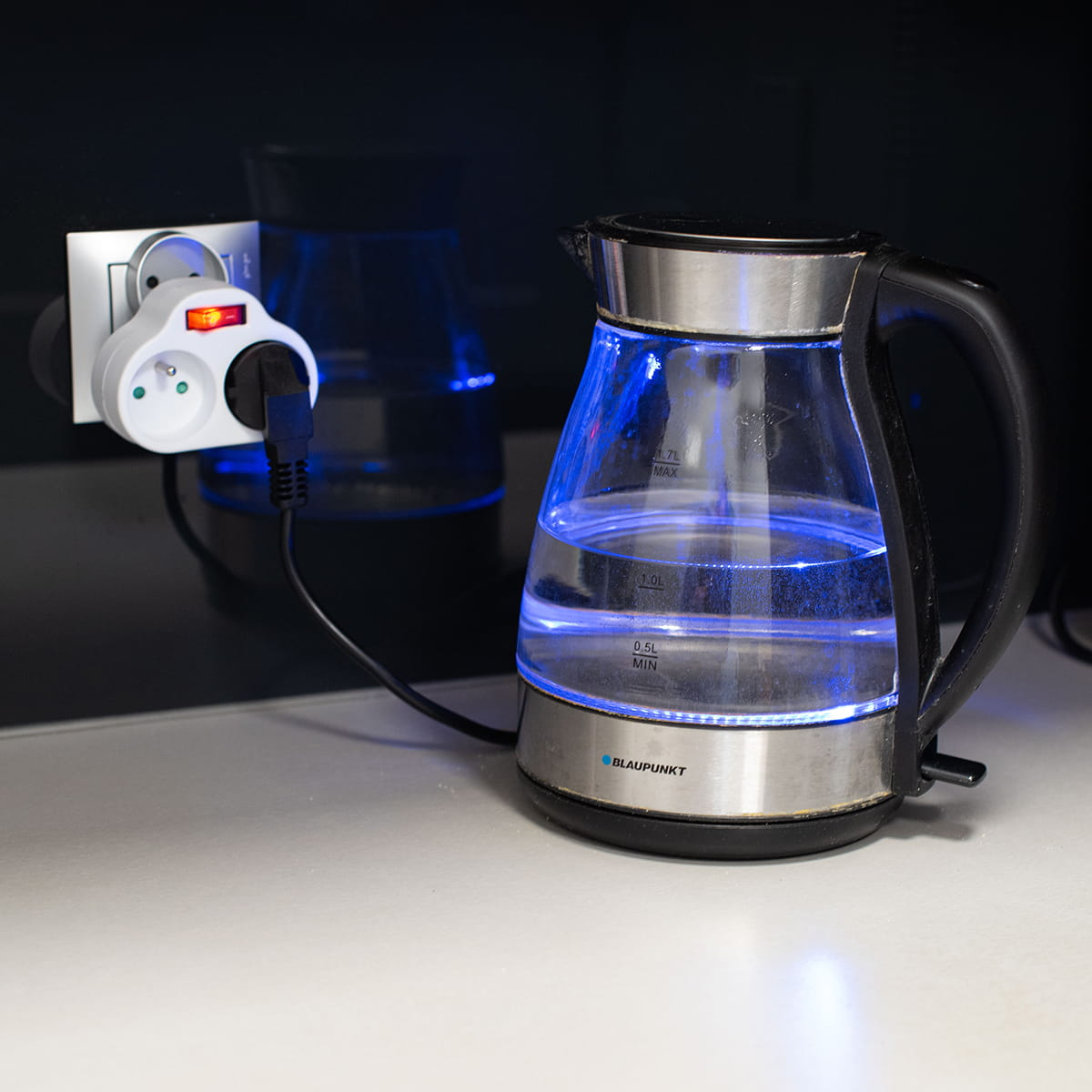 Szklany czajnik elektryczny z niebieskim światłem, podłączony do adaptera na białej powierzchni.