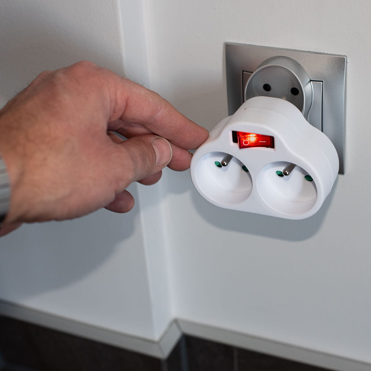 Ręka dotykająca biały adapter z czerwonym przełącznikiem wpięty do gniazdka.