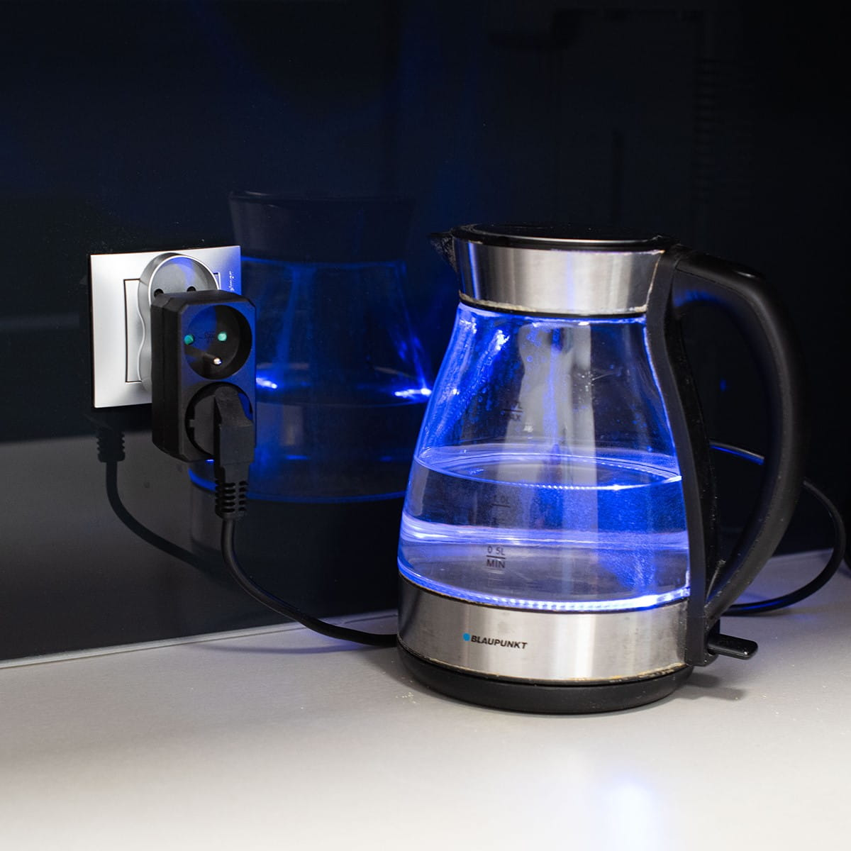 Przezroczysty czajnik elektryczny ze srebrną podstawą i uchwytem jest podłączony do gniazdka ściennego.