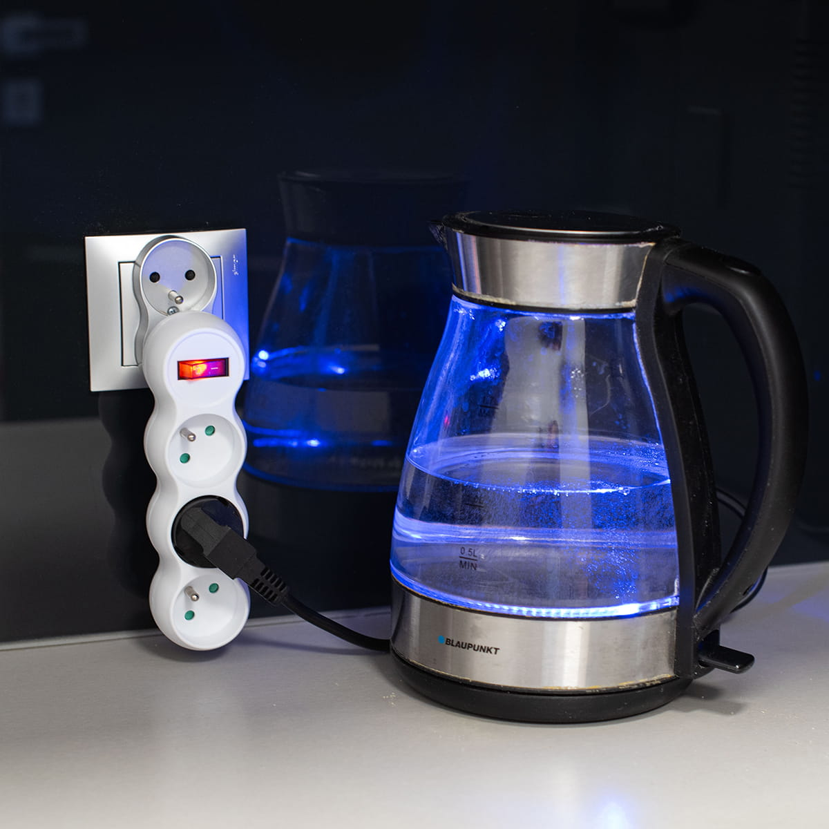Przezroczysty czajnik elektryczny ze srebrną podstawą, podłączony do białej listwy zasilającej na szarym blacie.