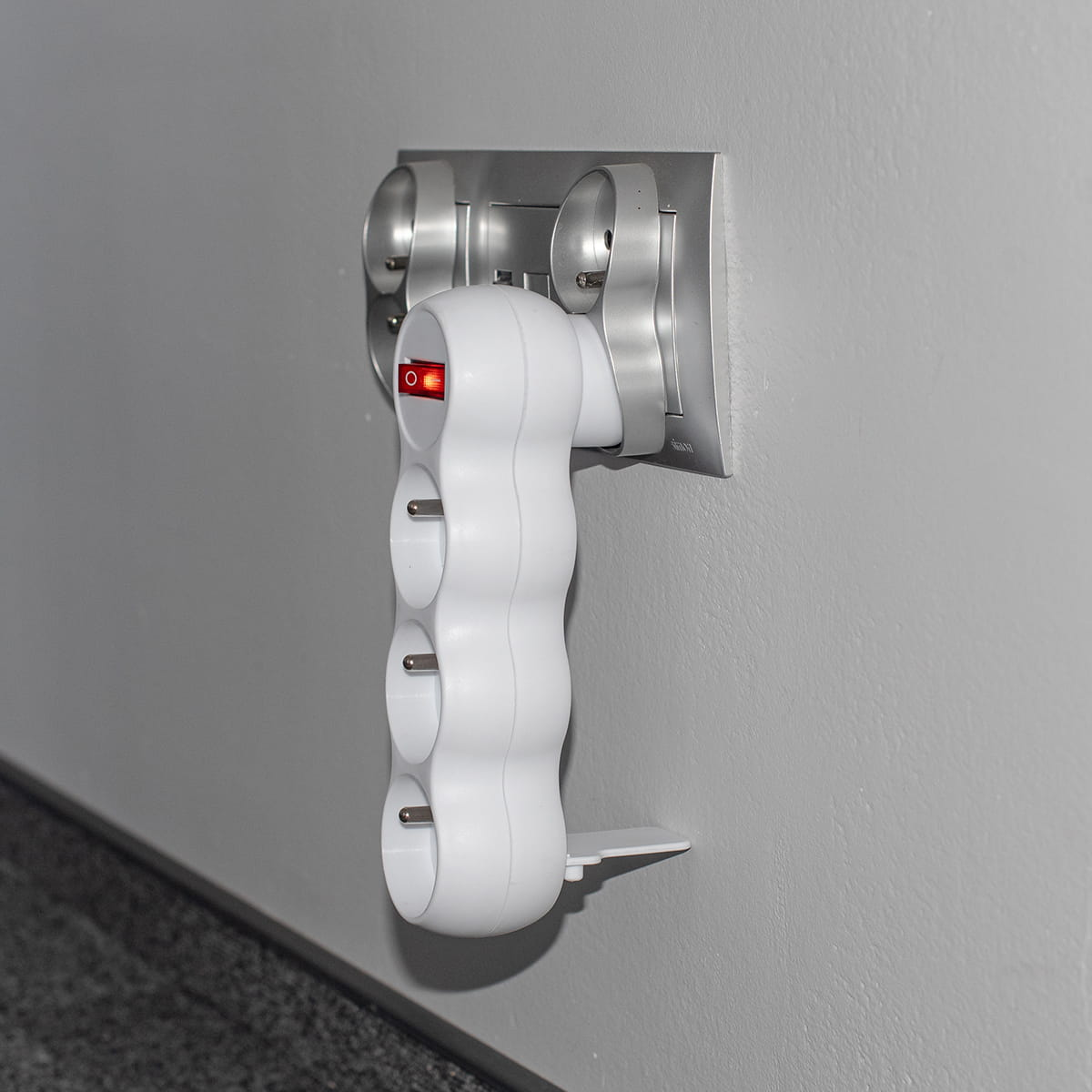 Biała listwa zasilająca z trzema gniazdami jest podłączona do gniazdka ściennego na szarej ścianie.