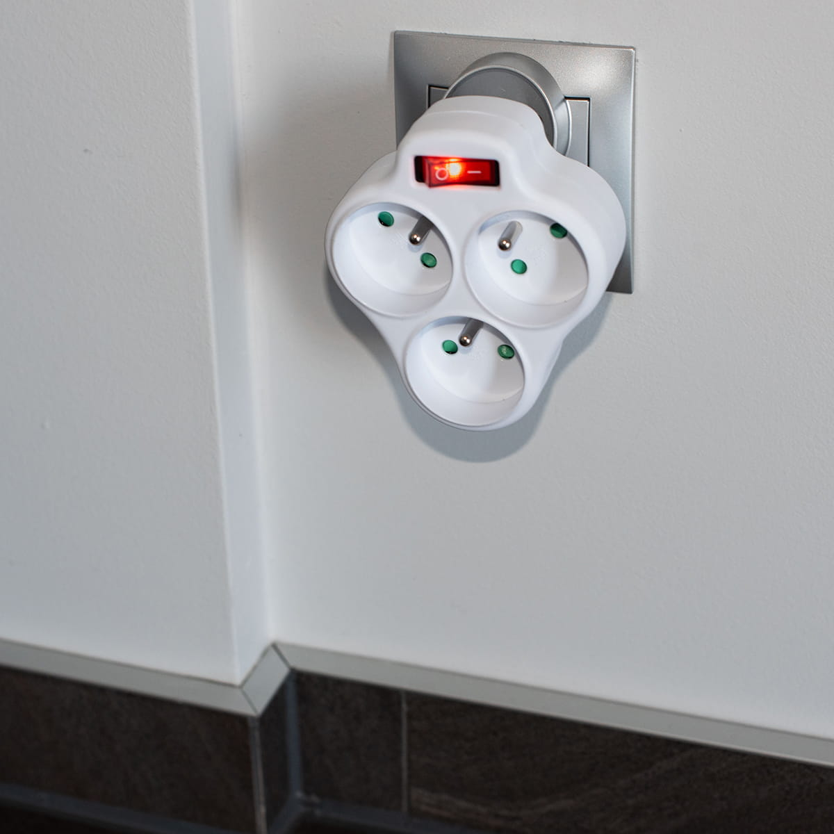 Białe potrójne gniazdo wpięte w gniazdo ścienne, z czerwonym włącznikiem.