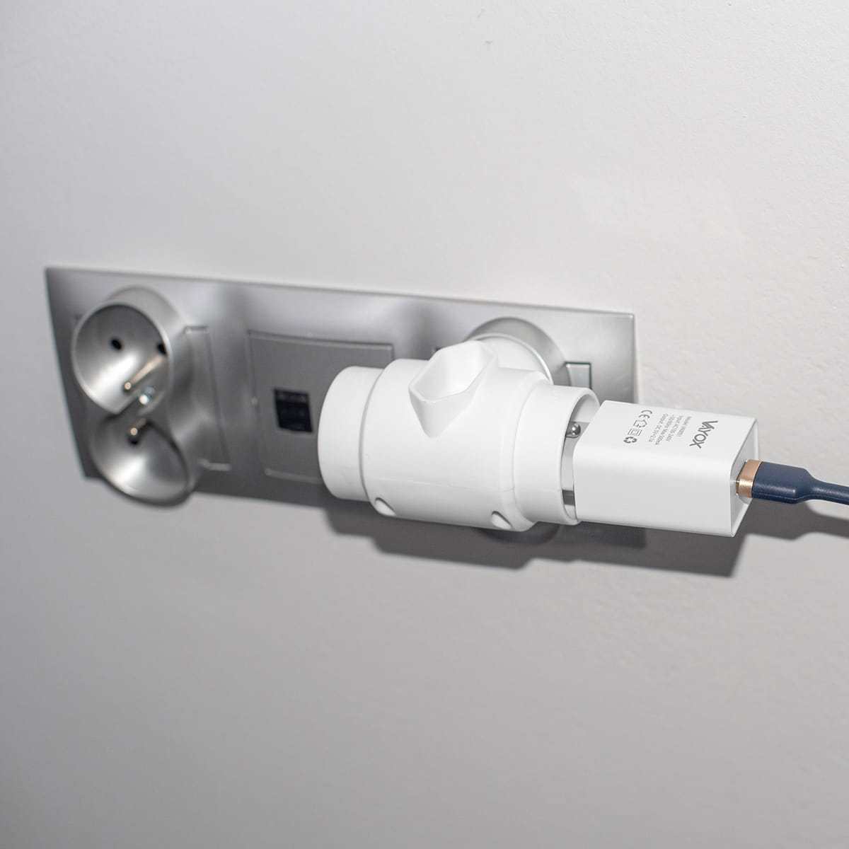 Biały adapter zasilania podłączony do srebrnego gniazda ściennego. Podłączona jest ładowarka do telefonu.