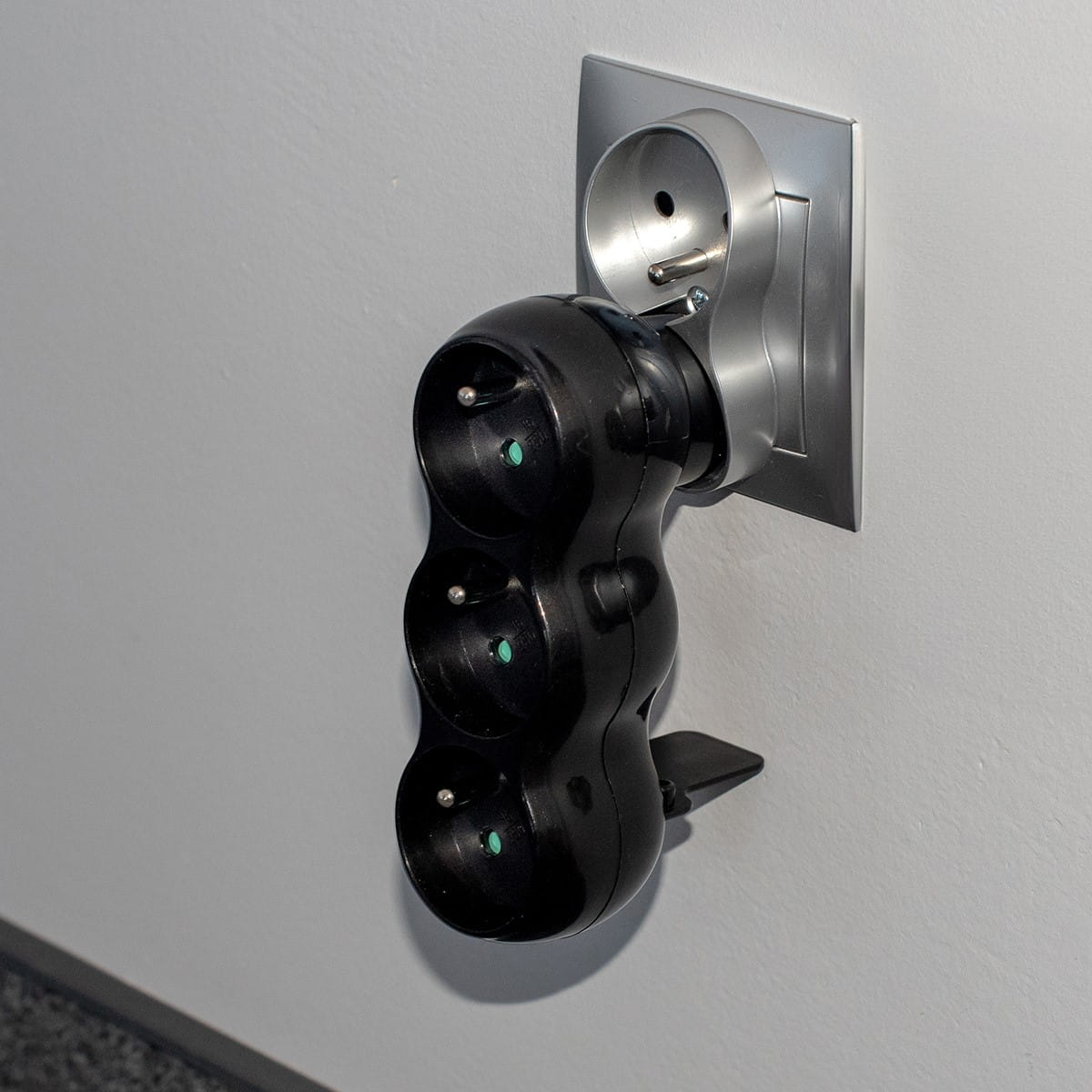 Czarny potrójny adapter zasilania podłączony do gniazdka ściennego. Ściana jest szara. Zbliżenie.