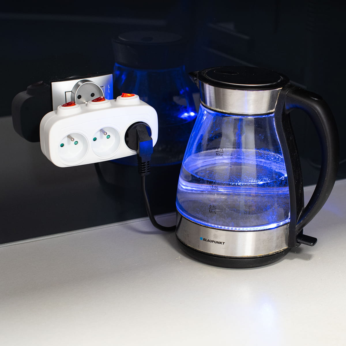 Biała listwa zasilająca z trzema gniazdami, podłączona do gniazdka ściennego. Czajnik jest podłączony i podświetlony.