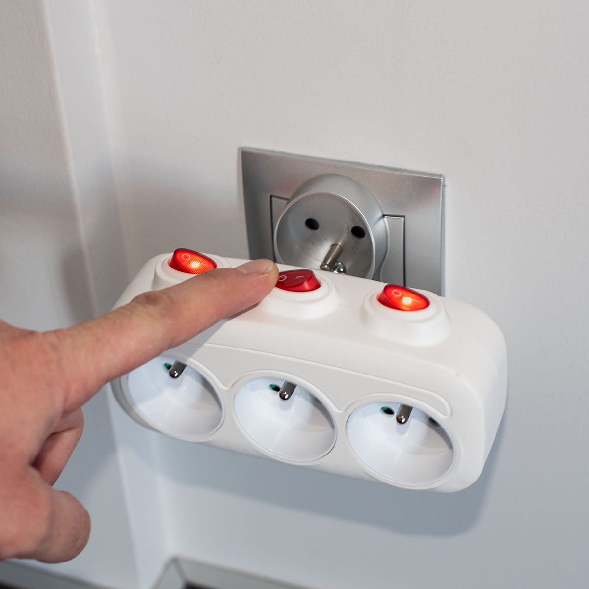 Ręka naciska czerwony przycisk na białej listwie zasilającej z trzema gniazdami. Gniazda są używane.