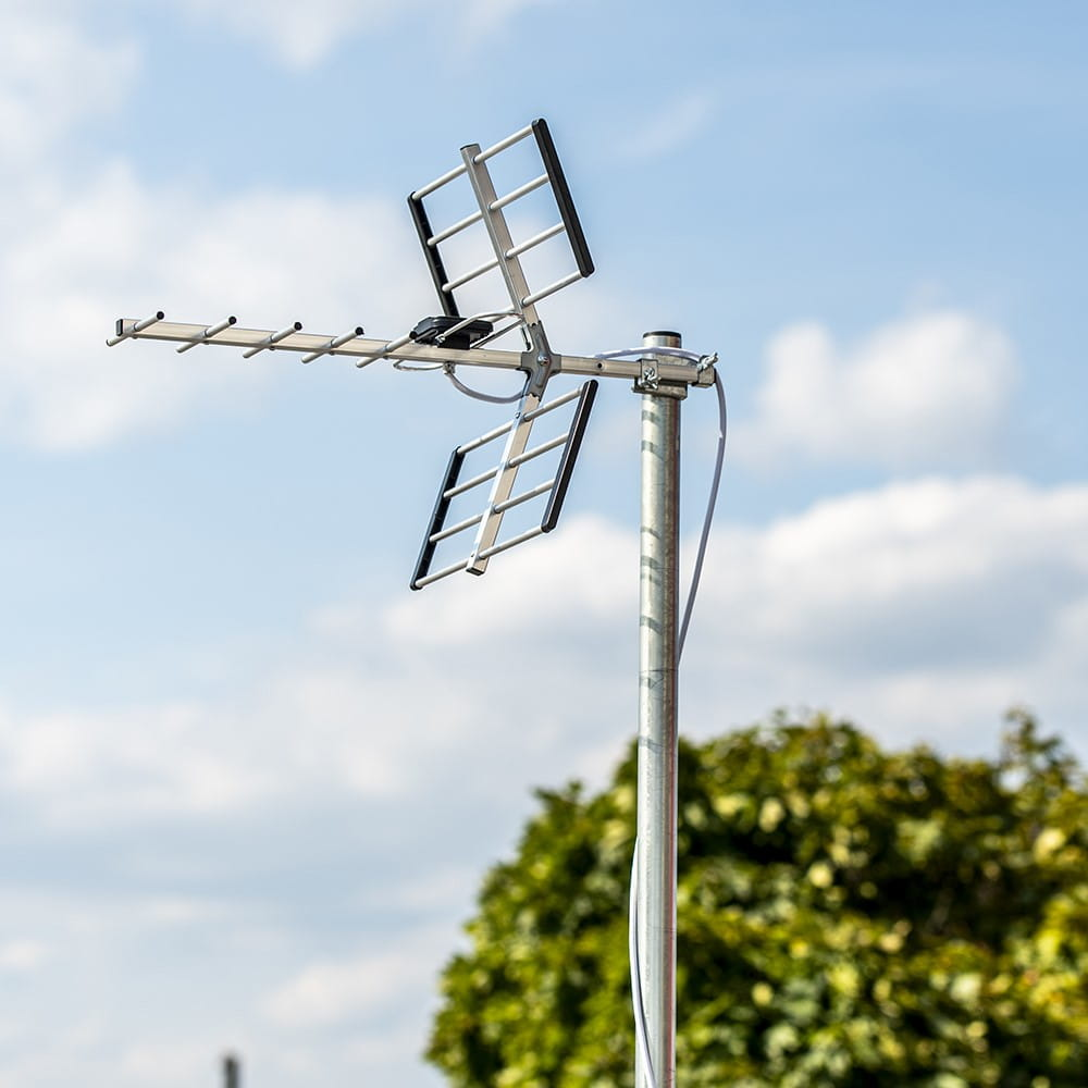 Antena na słupie na tle zachmurzonego błękitnego nieba i zielonych liści.