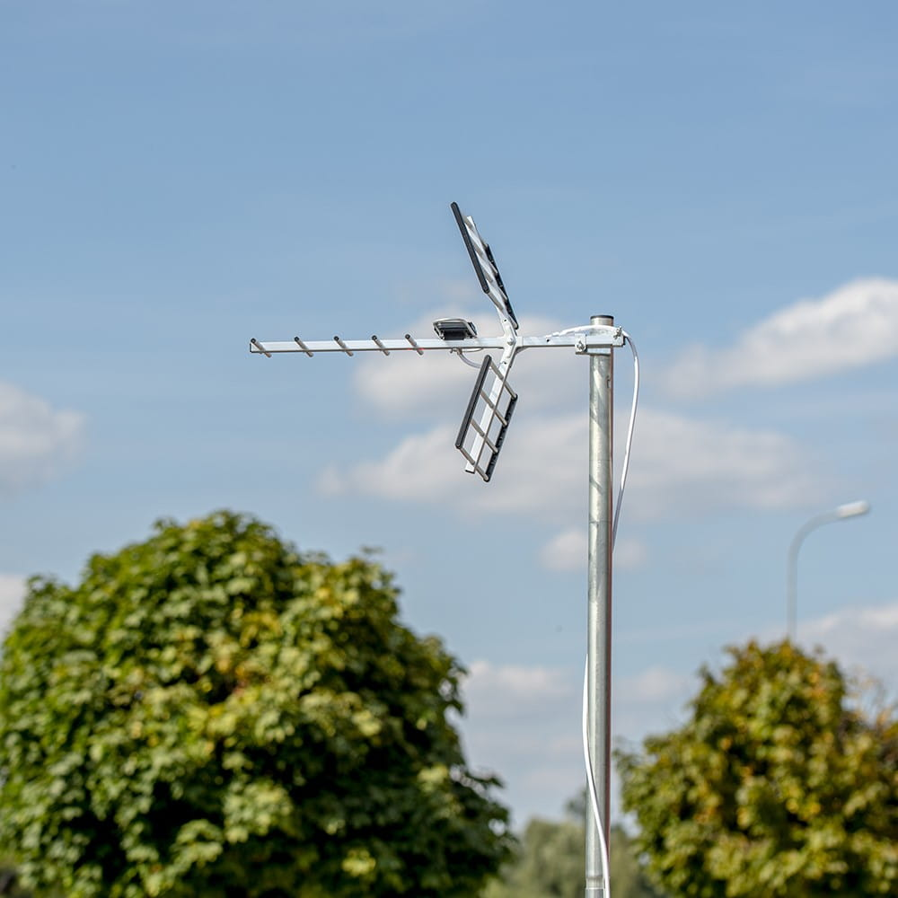 Antena telewizyjna na słupie, z niebieskim niebem w tle i zielonymi drzewami po bokach.