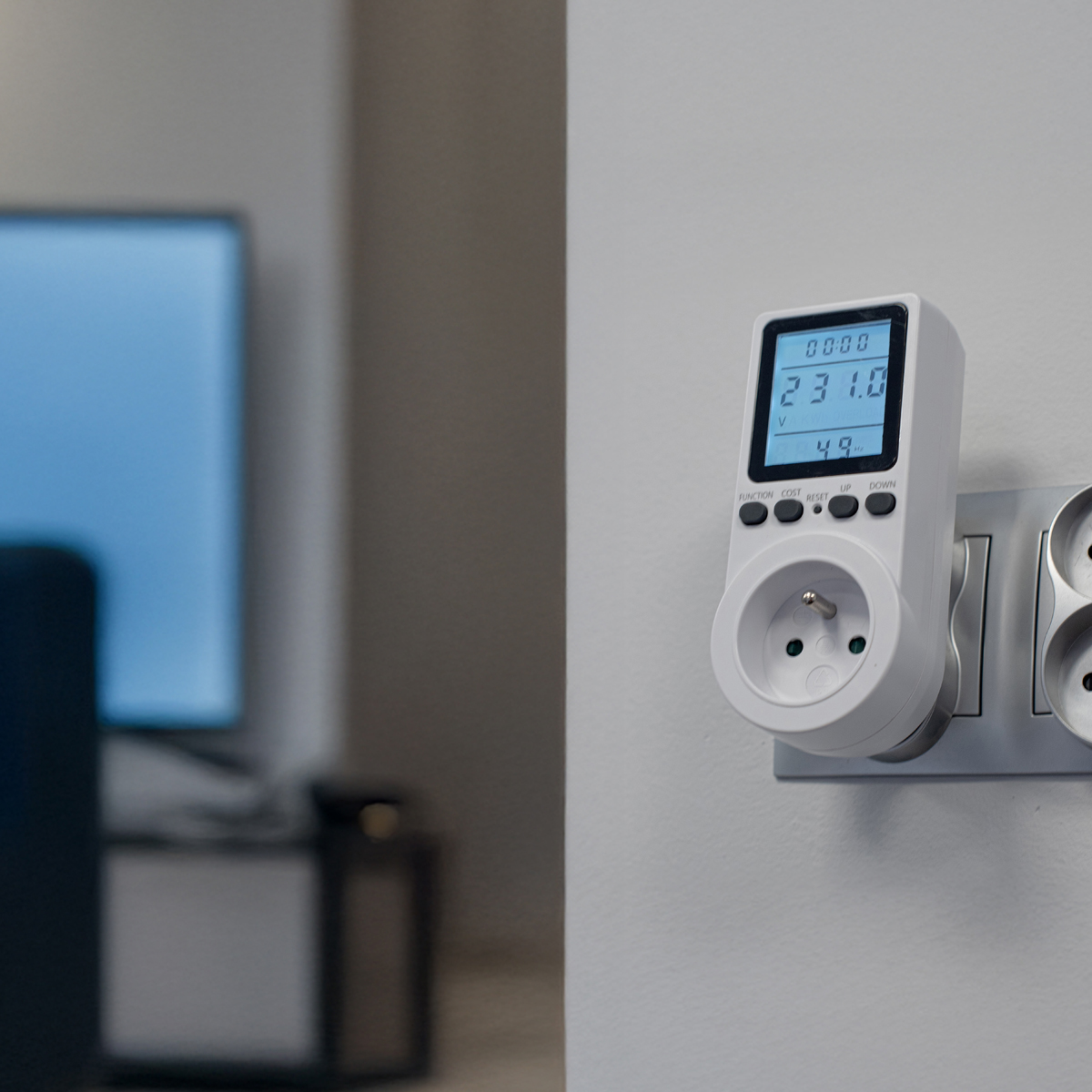Biały miernik energii w gniazdku, wyświetlający odczyty. W tle telewizor.