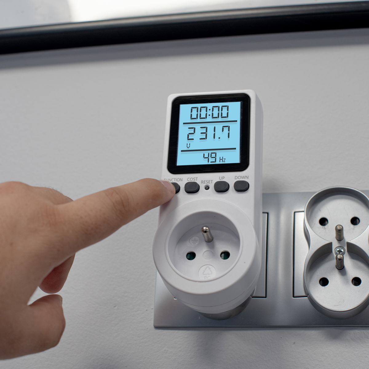 Ręka dotyka przycisku na białym mierniku mocy podłączonym do gniazdka ściennego.