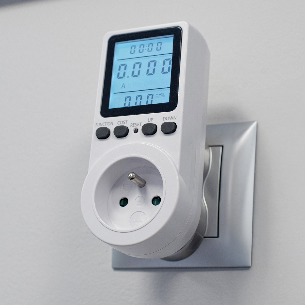 Biały miernik mocy wpięty w gniazdko ścienne, z danymi wyświetlanymi na ekranie.