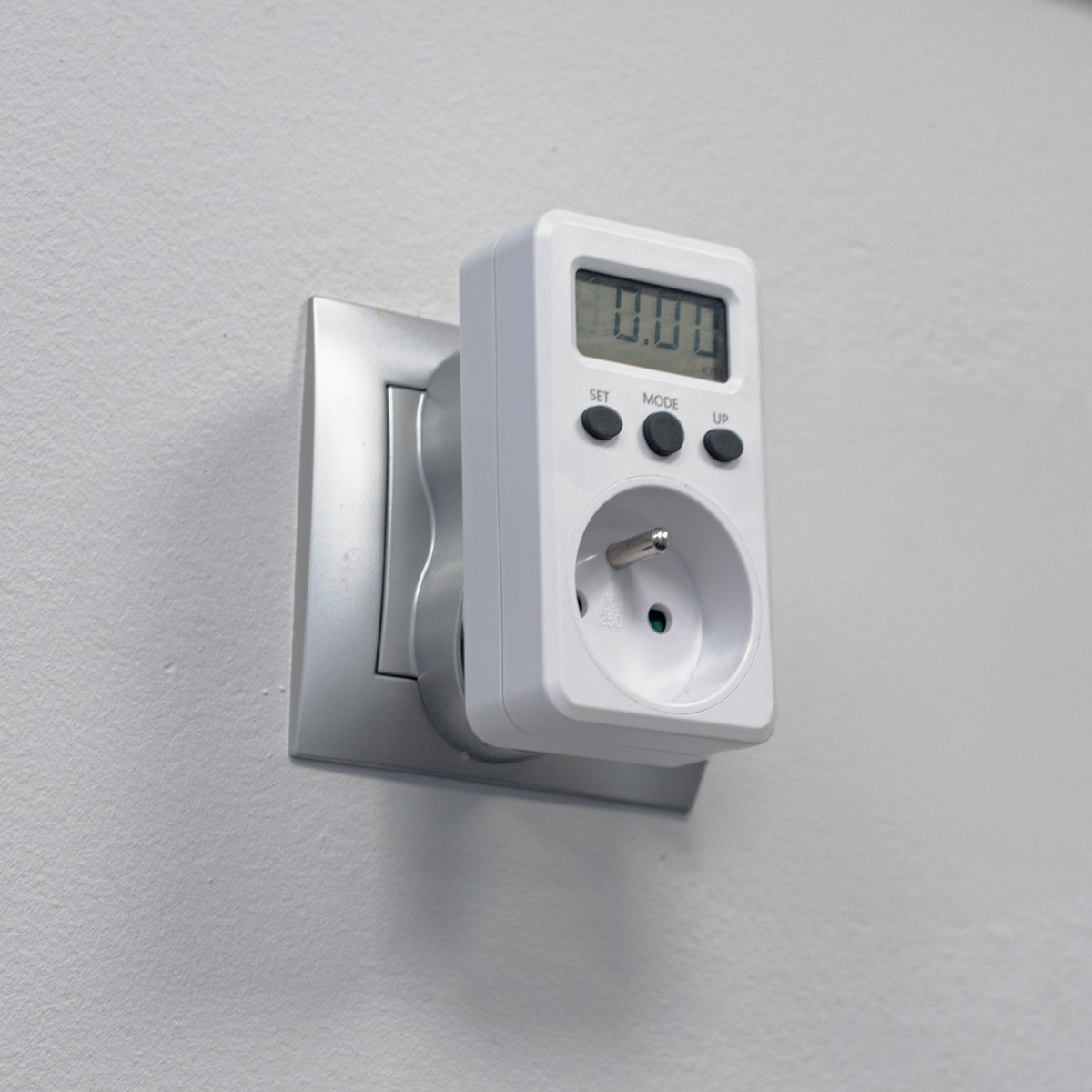 Biały miernik mocy podłączony do srebrnego gniazda ściennego.