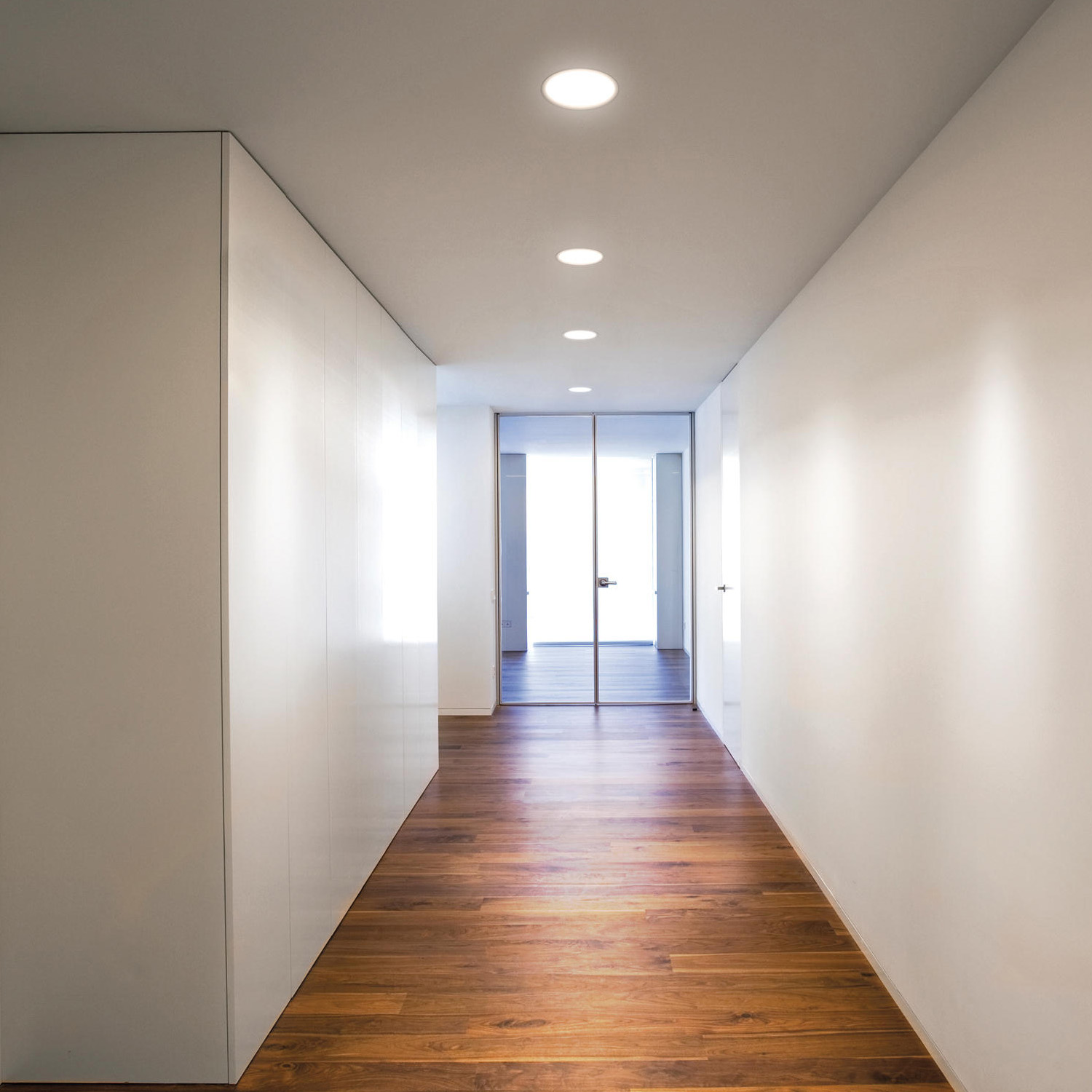 Un pasillo largo y estrecho con suelo de madera, paredes blancas y luces empotradas en el techo. Hay una puerta al final.