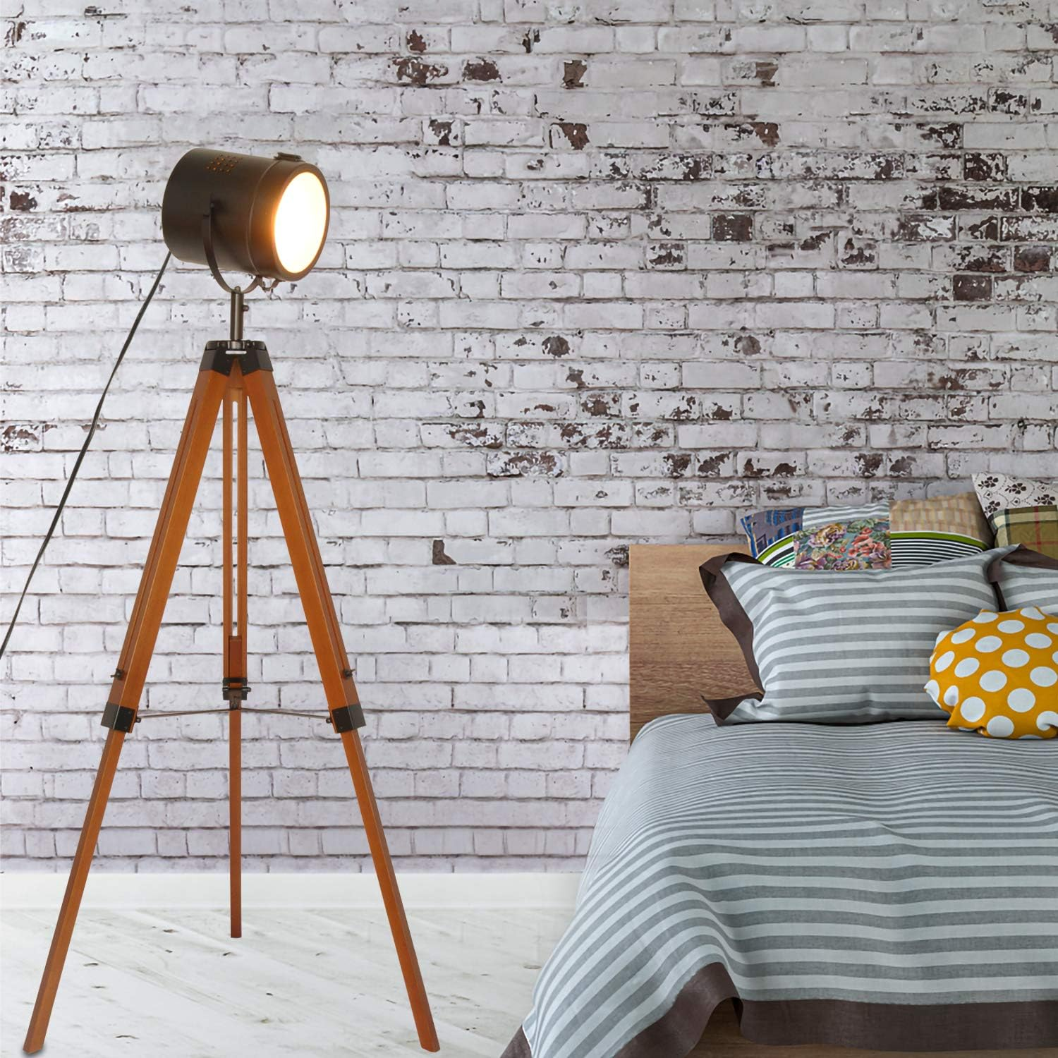 Una lámpara trípode de madera con foco, contra una pared de ladrillo y cama.