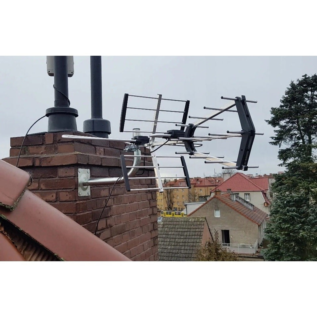 Antena zamontowana na ceglanym kominie. Tło przedstawia krajobraz miejski pod zachmurzonym niebem.