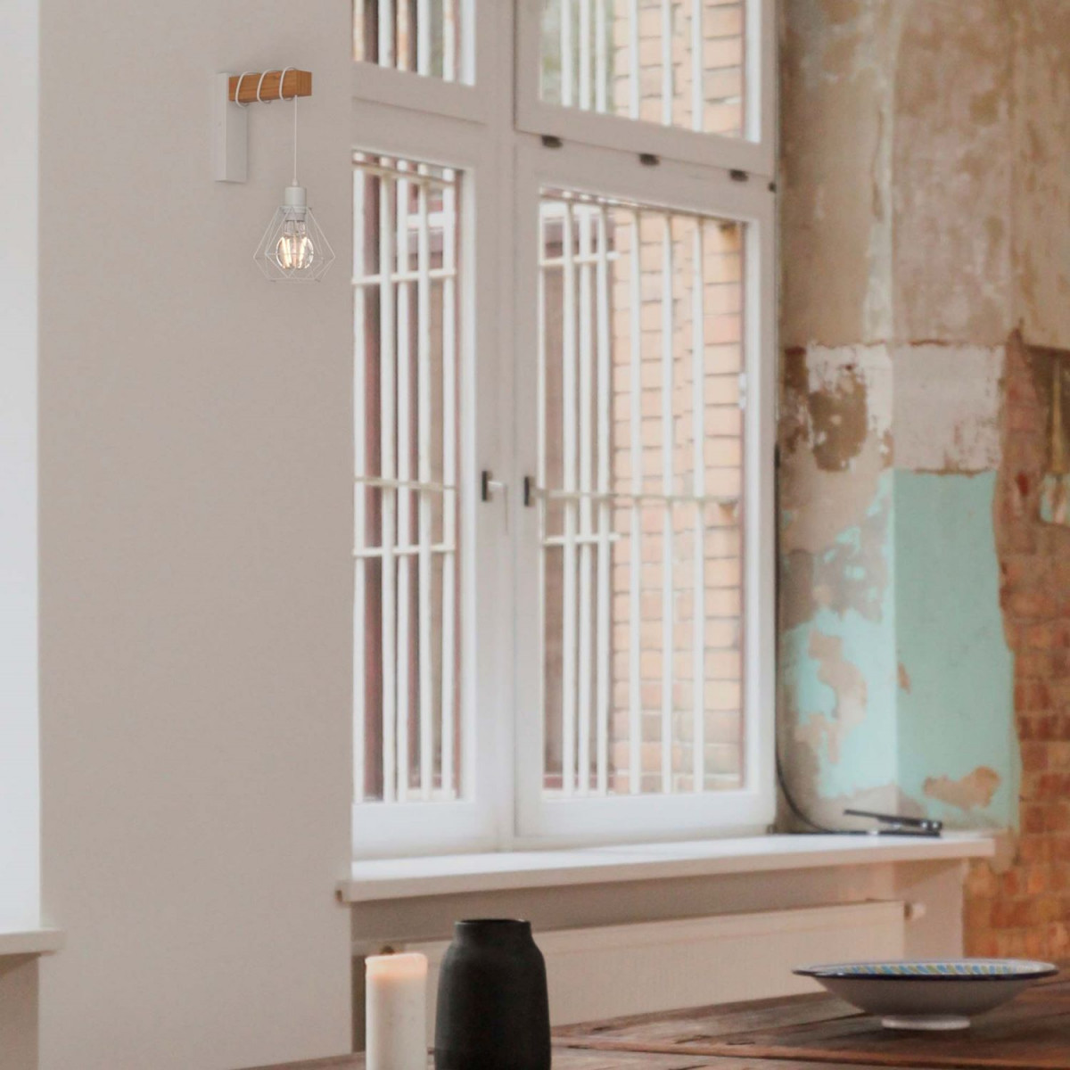 Una moderna lámpara junto a una ventana, pared blanca y mesa de madera. Un jarrón y un cuenco en la mesa.