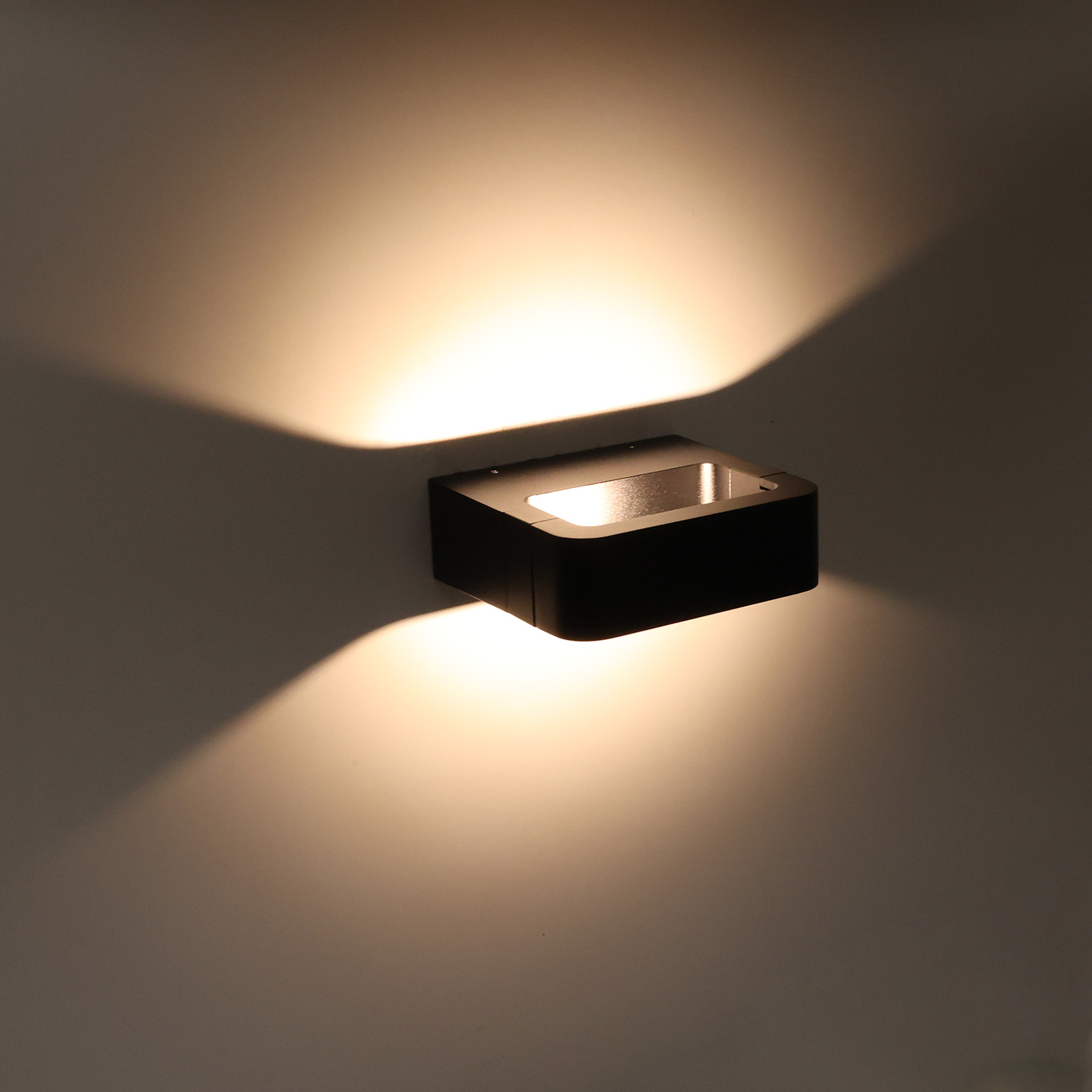 Una lámpara de pared rectangular negra emitiendo luz sobre una pared gris. La luz tiene dirección descendente.