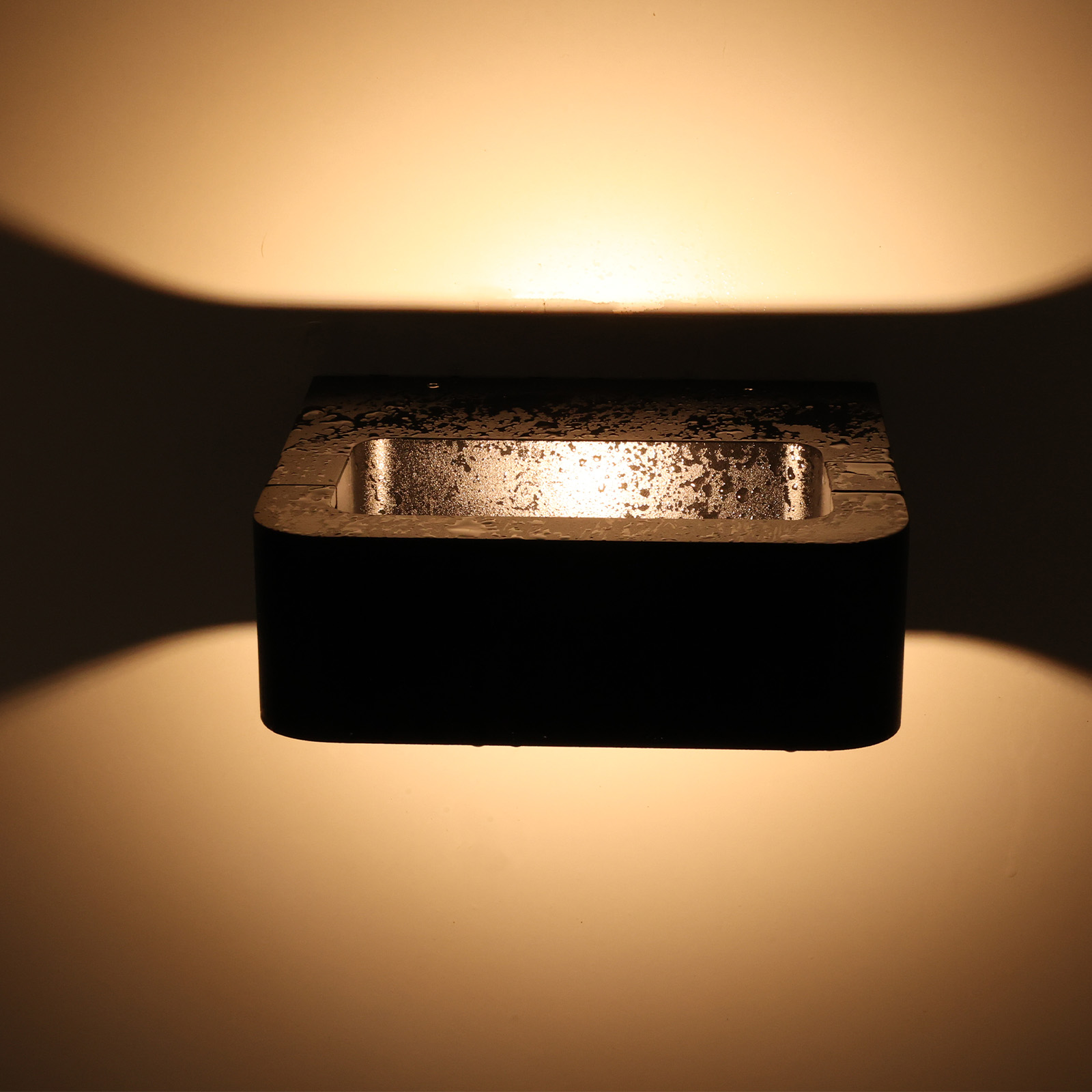 Una lámpara de pared rectangular negra está encendida, proyectando un brillo cálido en la pared. Gotas de agua visibles.
