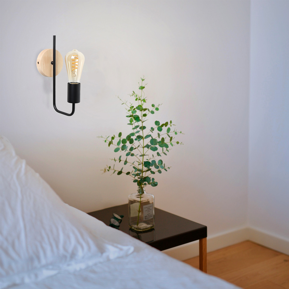 Una escena de dormitorio con una lámpara de pared negra con bombilla, una cama y una planta en un jarrón sobre una mesita de noche.