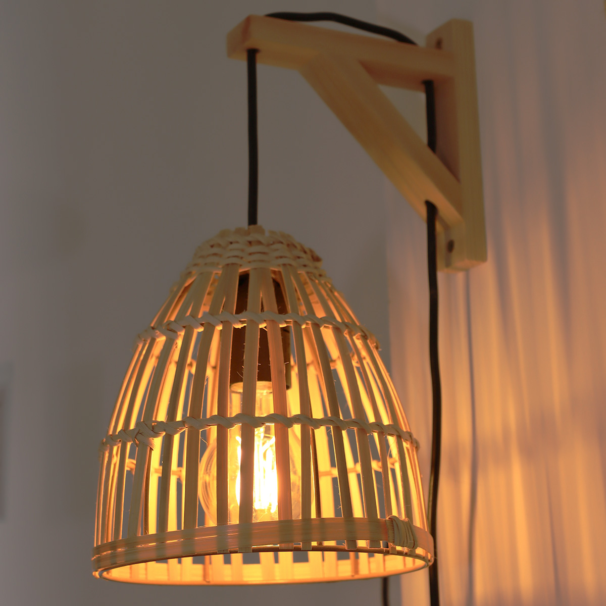 Una lámpara de pared con pantalla de mimbre, soporte de madera, cable negro y una bombilla encendida.