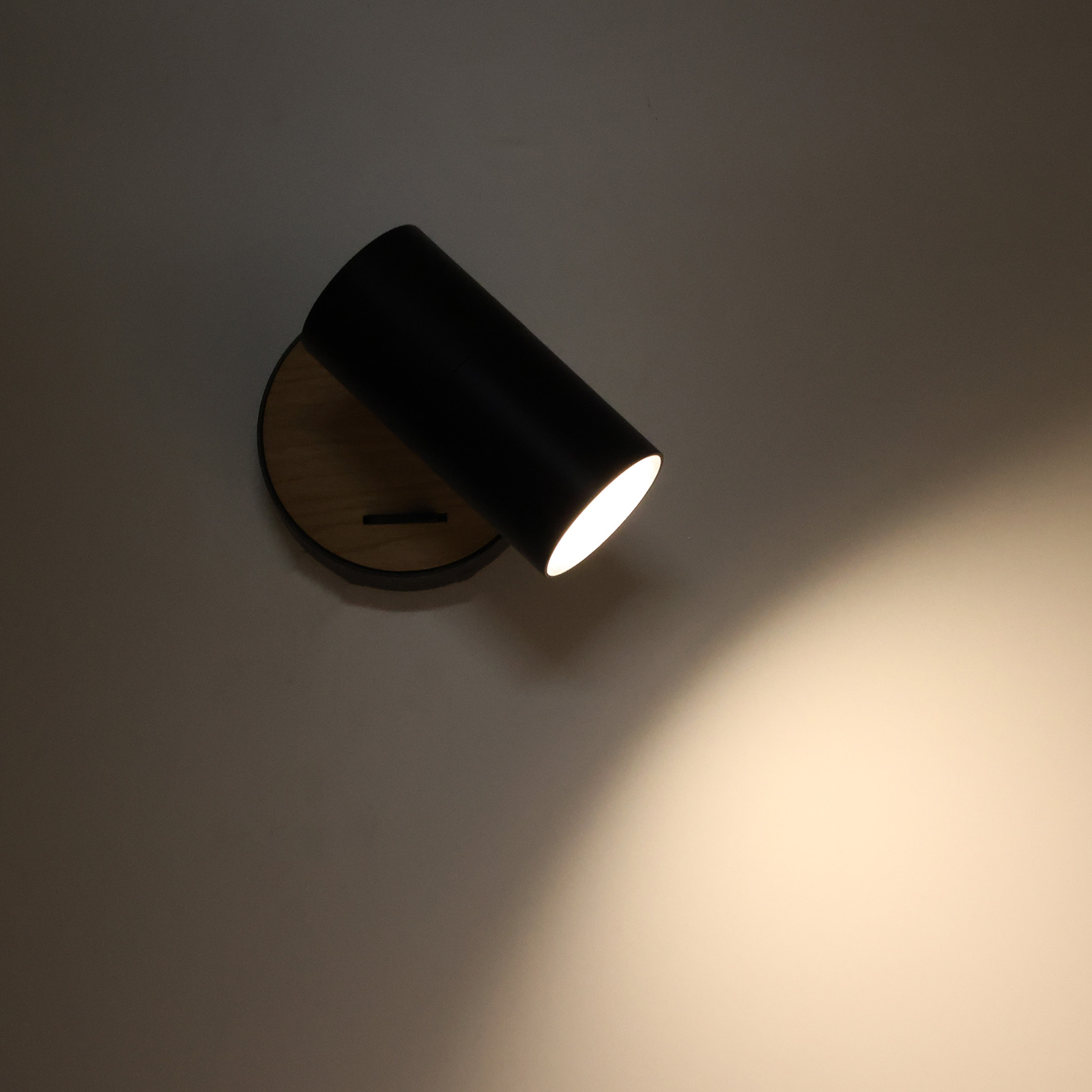 Foco negro sobre base de madera, inclinado hacia abajo, iluminando un punto en la pared de abajo.