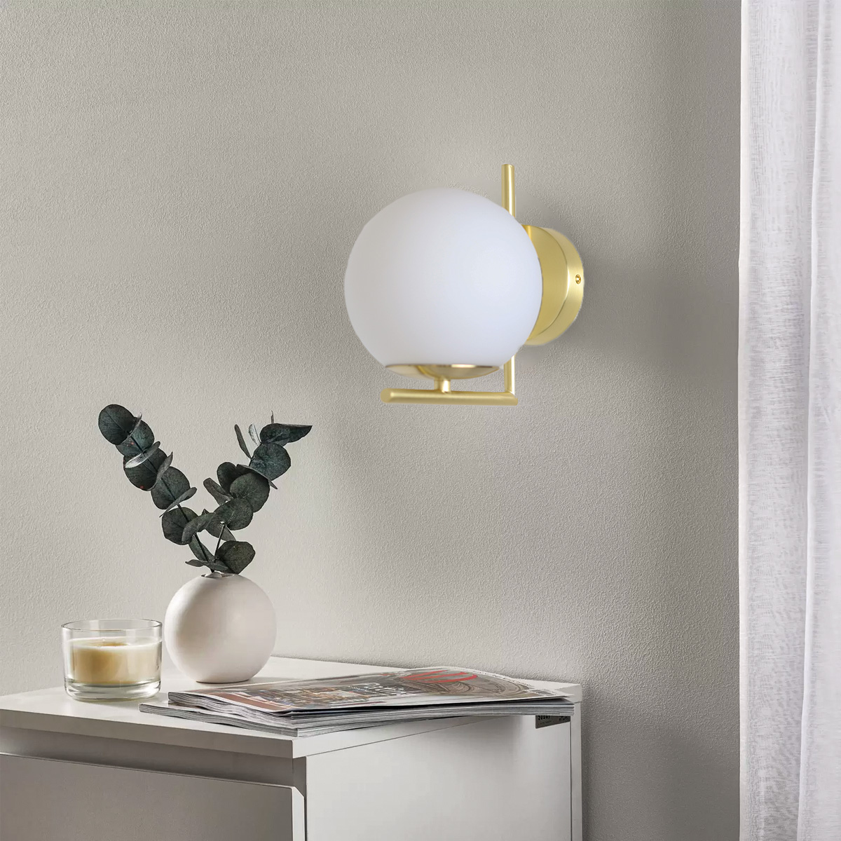 Una lámpara montada en la pared con una pantalla de orbe blanca. Está en una pared junto a un jarrón, una vela y una revista.