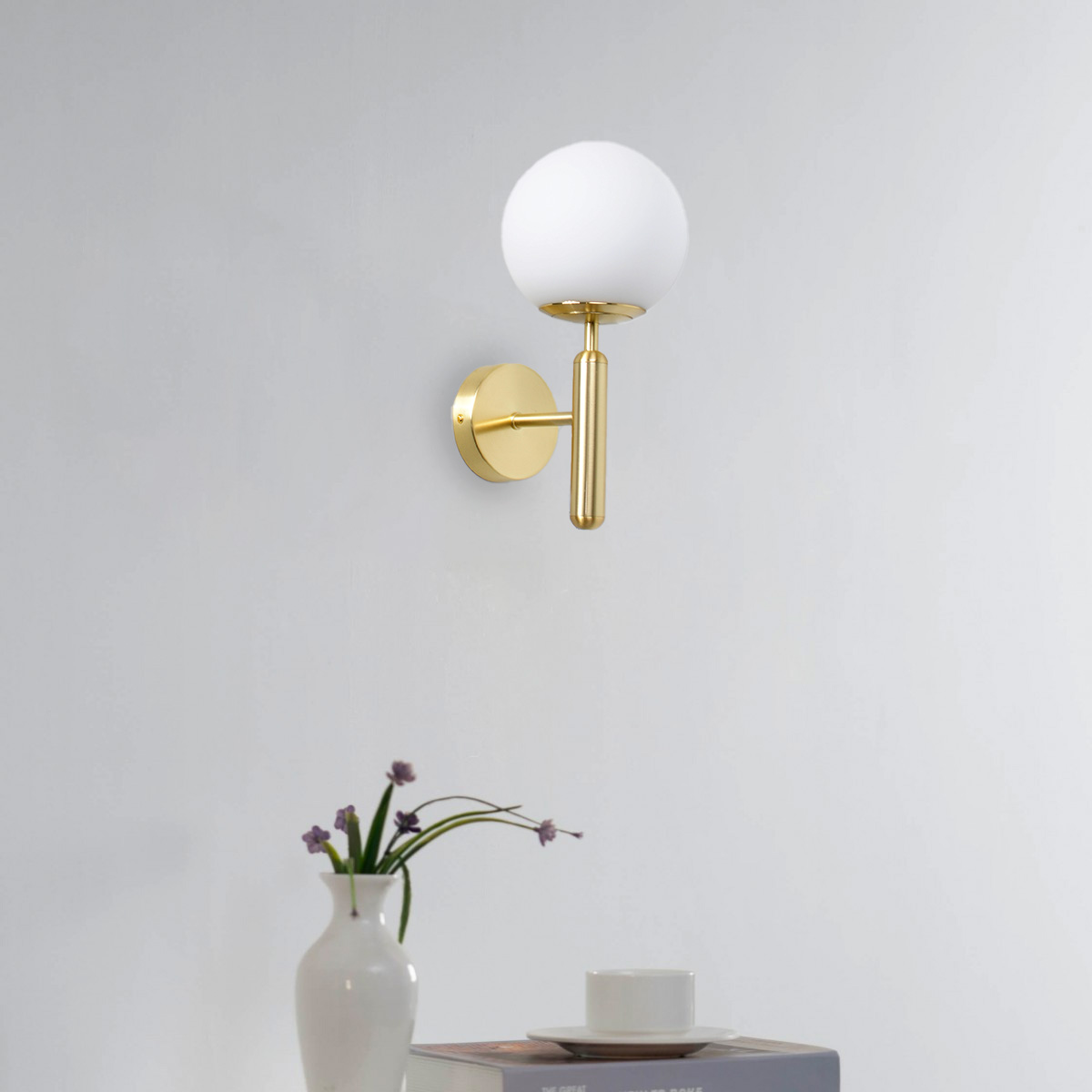Una lámpara de pared dorada con una bombilla blanca en forma de globo. Un jarrón con flores y libros debajo de la lámpara.