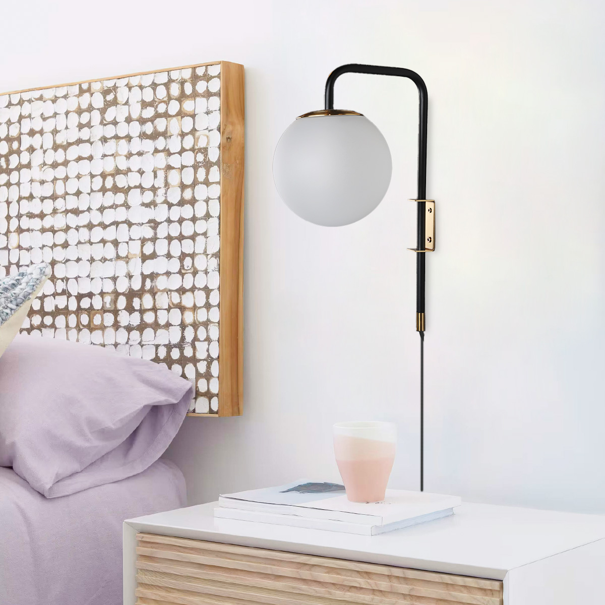 Lámpara de pared negra y dorada con bombilla globo blanca, junto a cama, taza rosa en mesa.