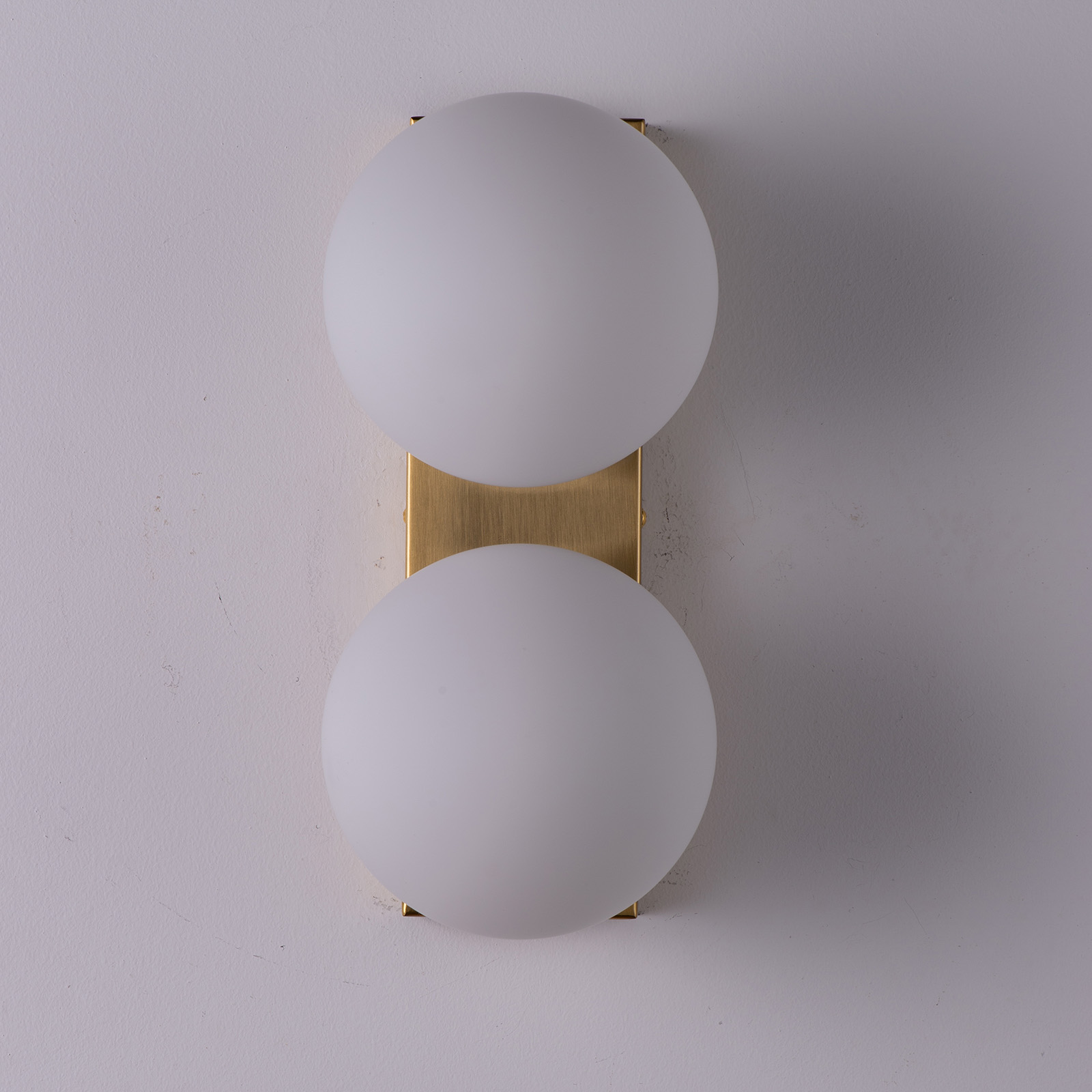 Dos bombillas blancas y redondas alineadas verticalmente en un soporte de pared rectangular dorado, contra una pared blanca.