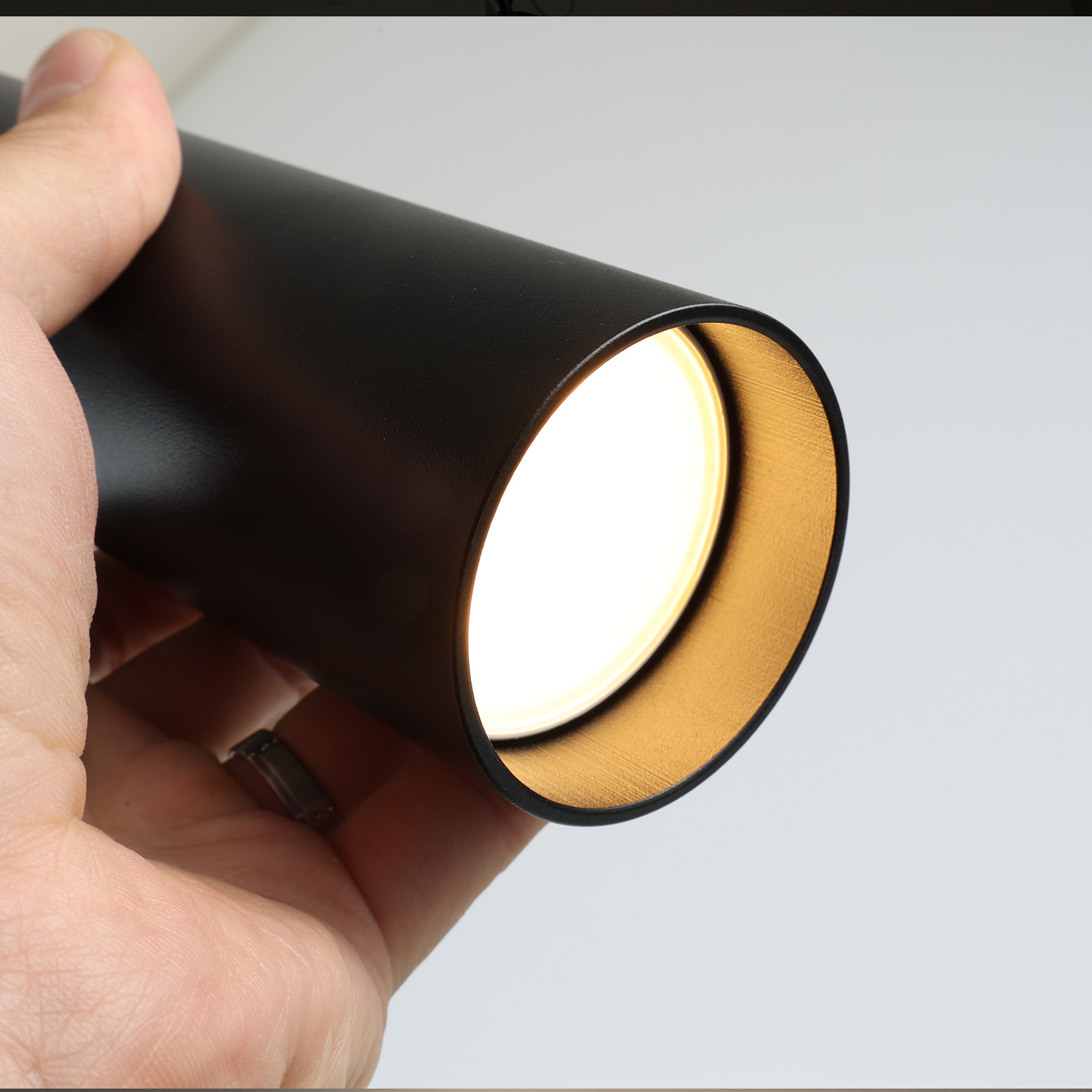 Una mano sosteniendo una luz cilíndrica negra, emitiendo luz brillante. Fondo blanco.