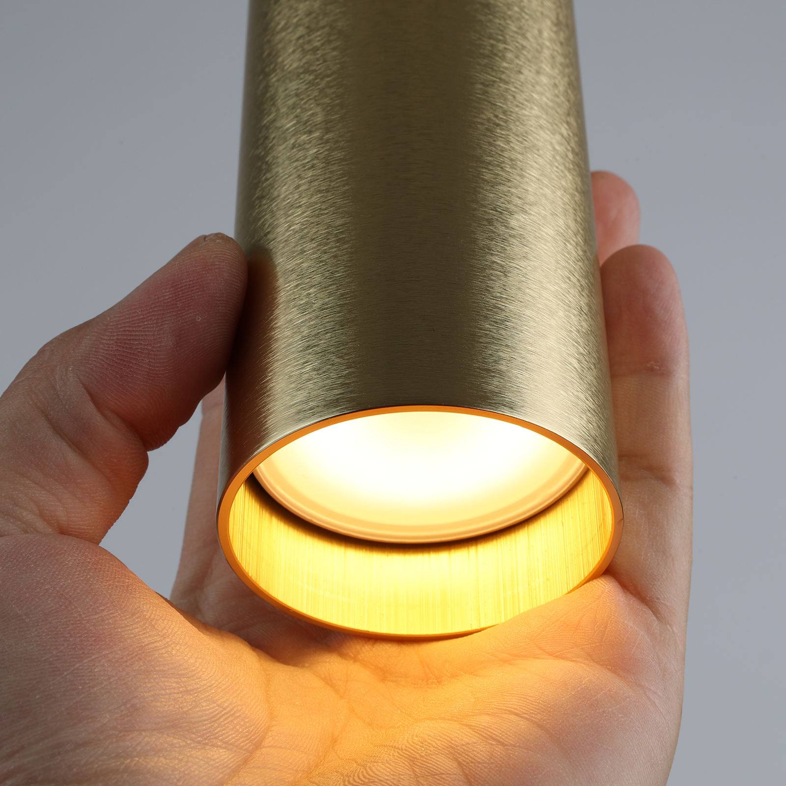 Una mano sostiene una lámpara colgante cilíndrica dorada, iluminada desde abajo.