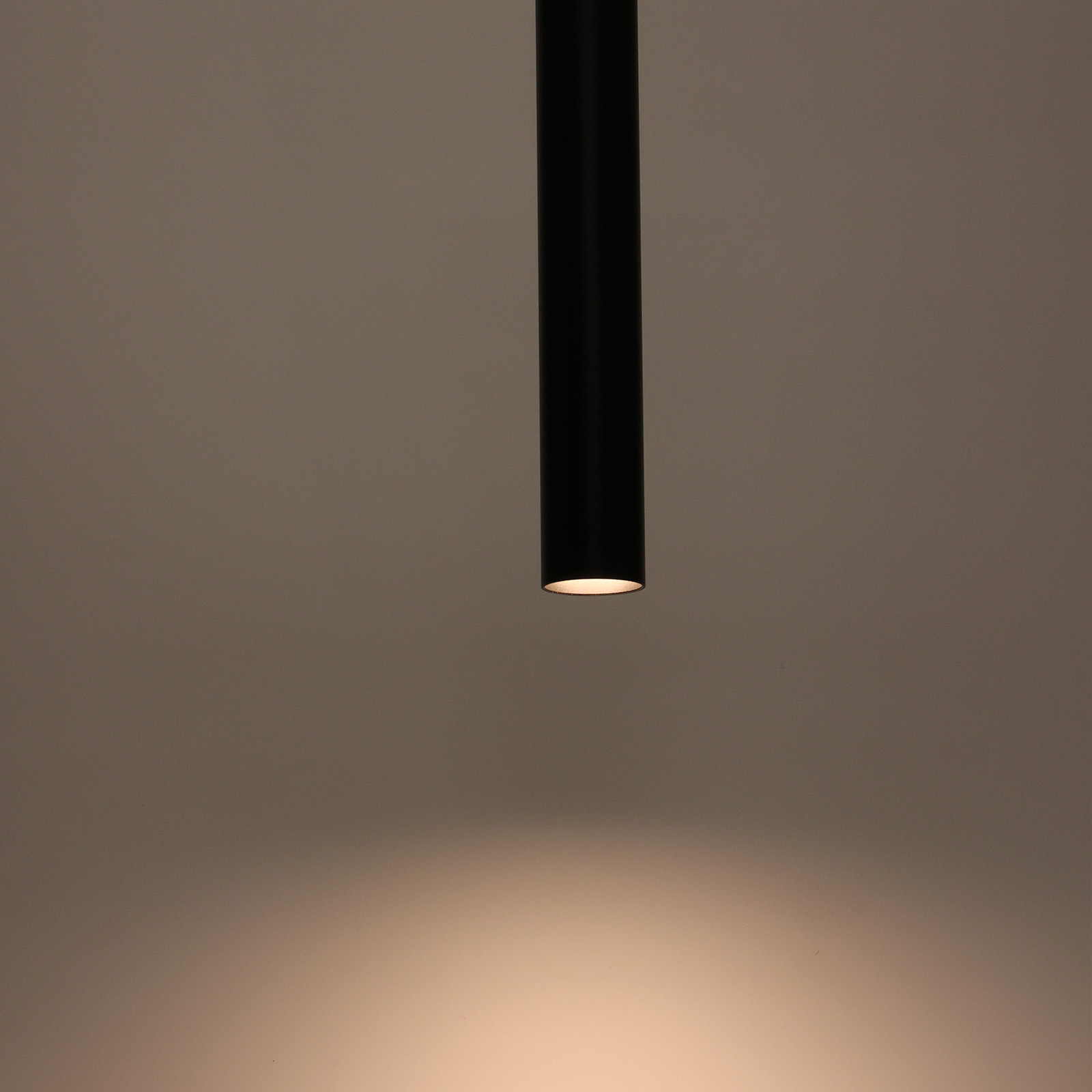 Lámpara colgante cilíndrica negra, iluminada desde abajo, colgando contra un fondo marrón.