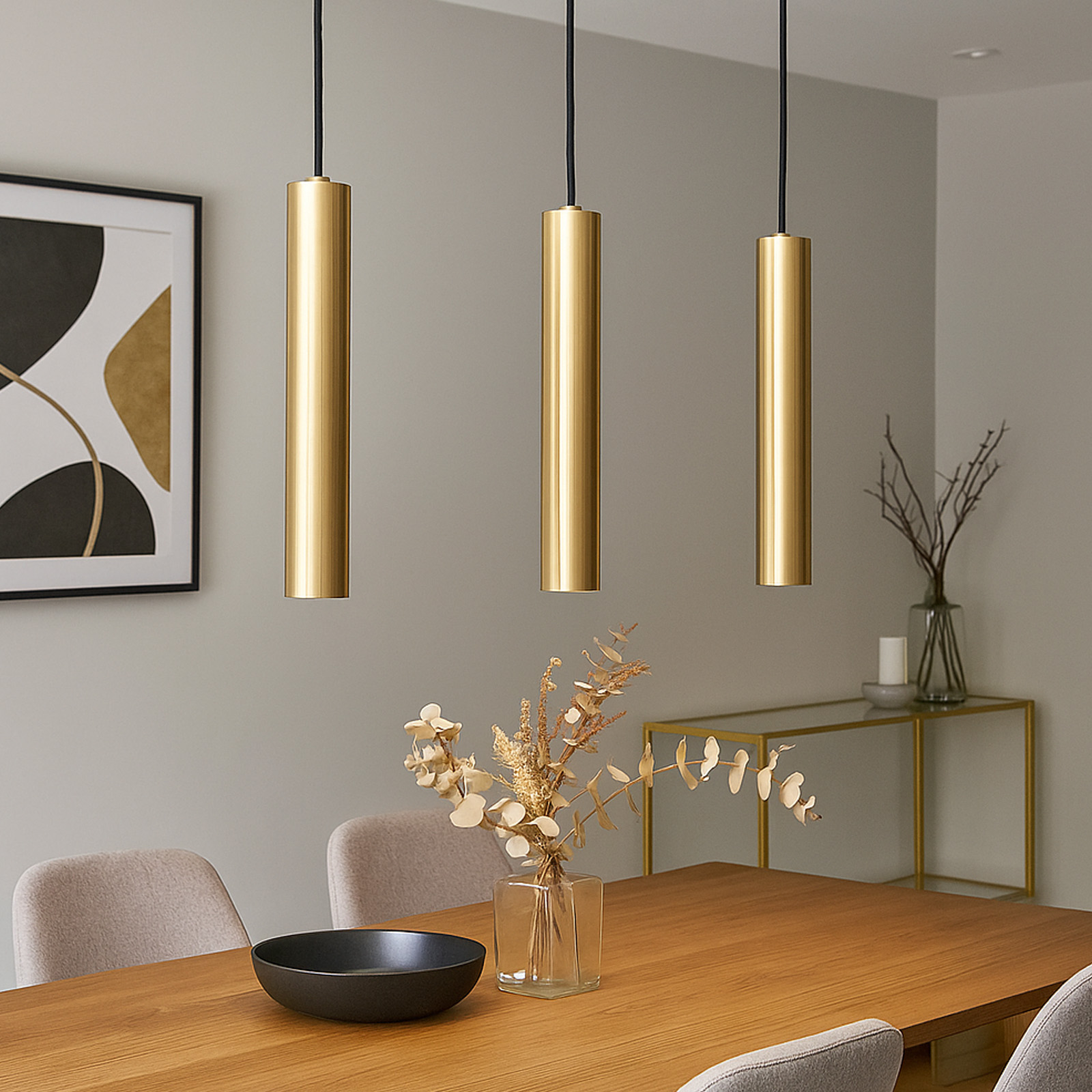 Tres luces colgantes cilíndricas doradas colgando sobre una mesa de madera, con decoración y un cuadro.