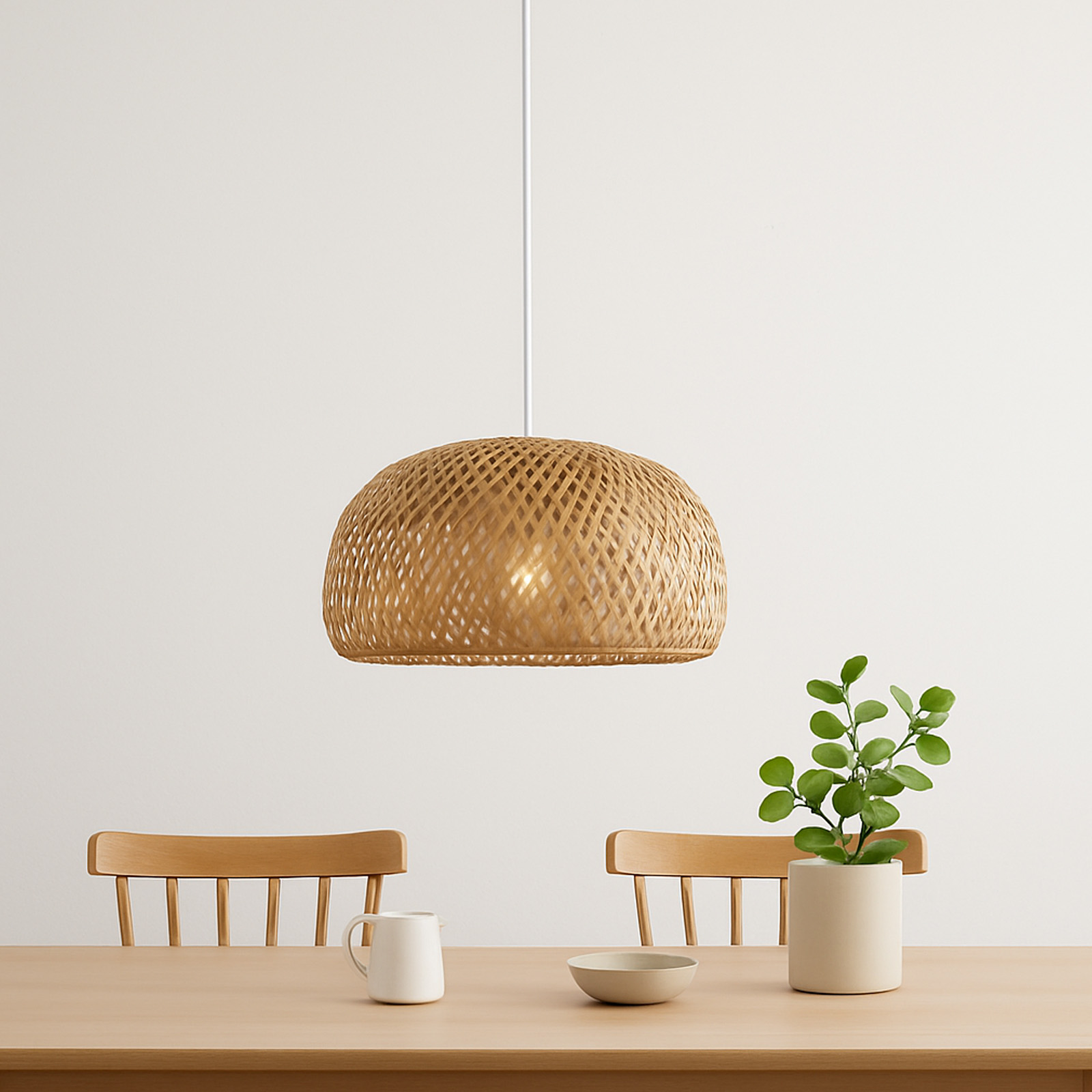 Una lámpara de bambú tejido colgando sobre una mesa de madera con una taza, un cuenco y una planta en un jarrón.