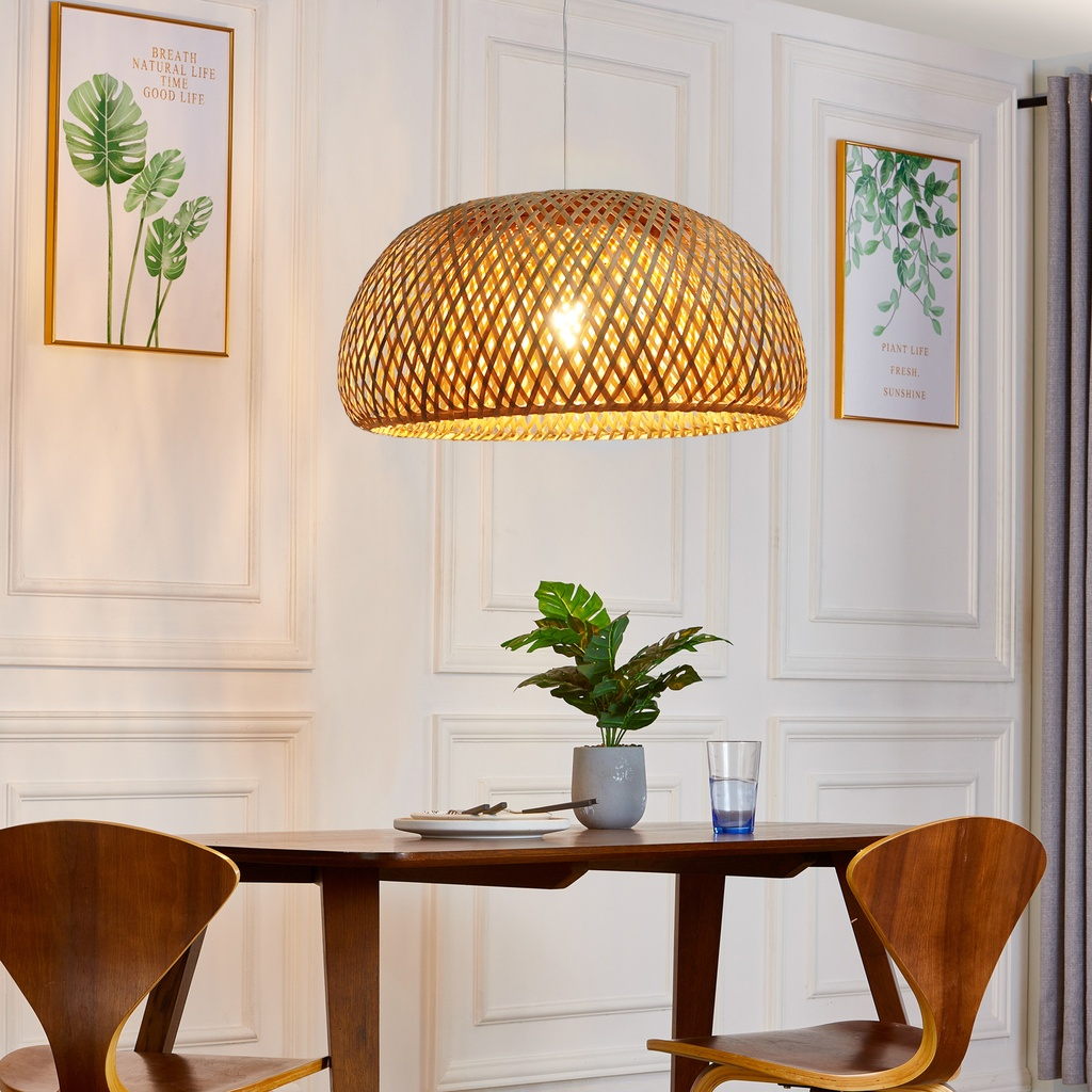 Una pantalla de lámpara de bambú tejido cuelga sobre una mesa de comedor de madera. Se ven obras de arte y una planta.