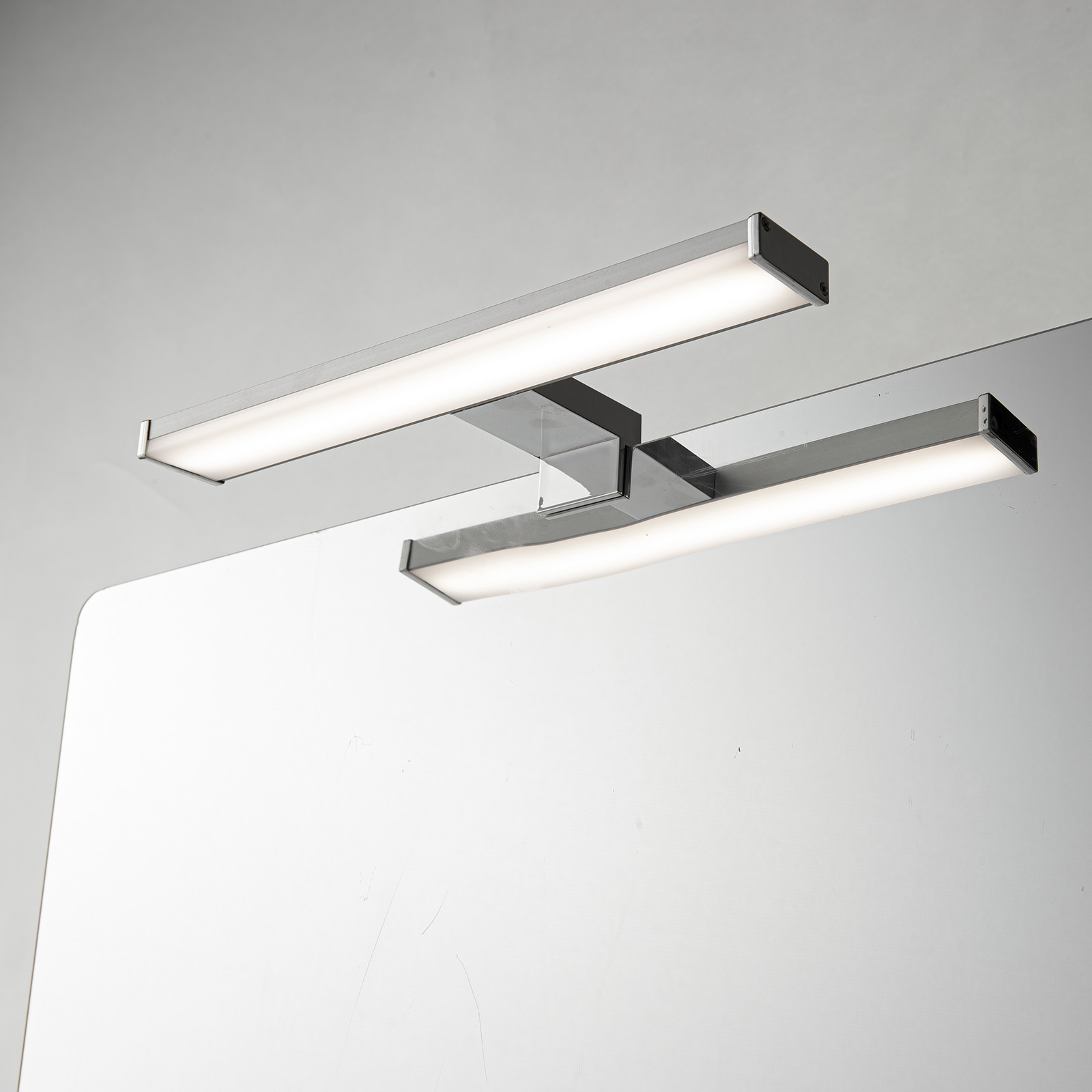Dos luces de tocador están montadas encima de un espejo. Las luces son plateadas y rectangulares, y el fondo es gris.